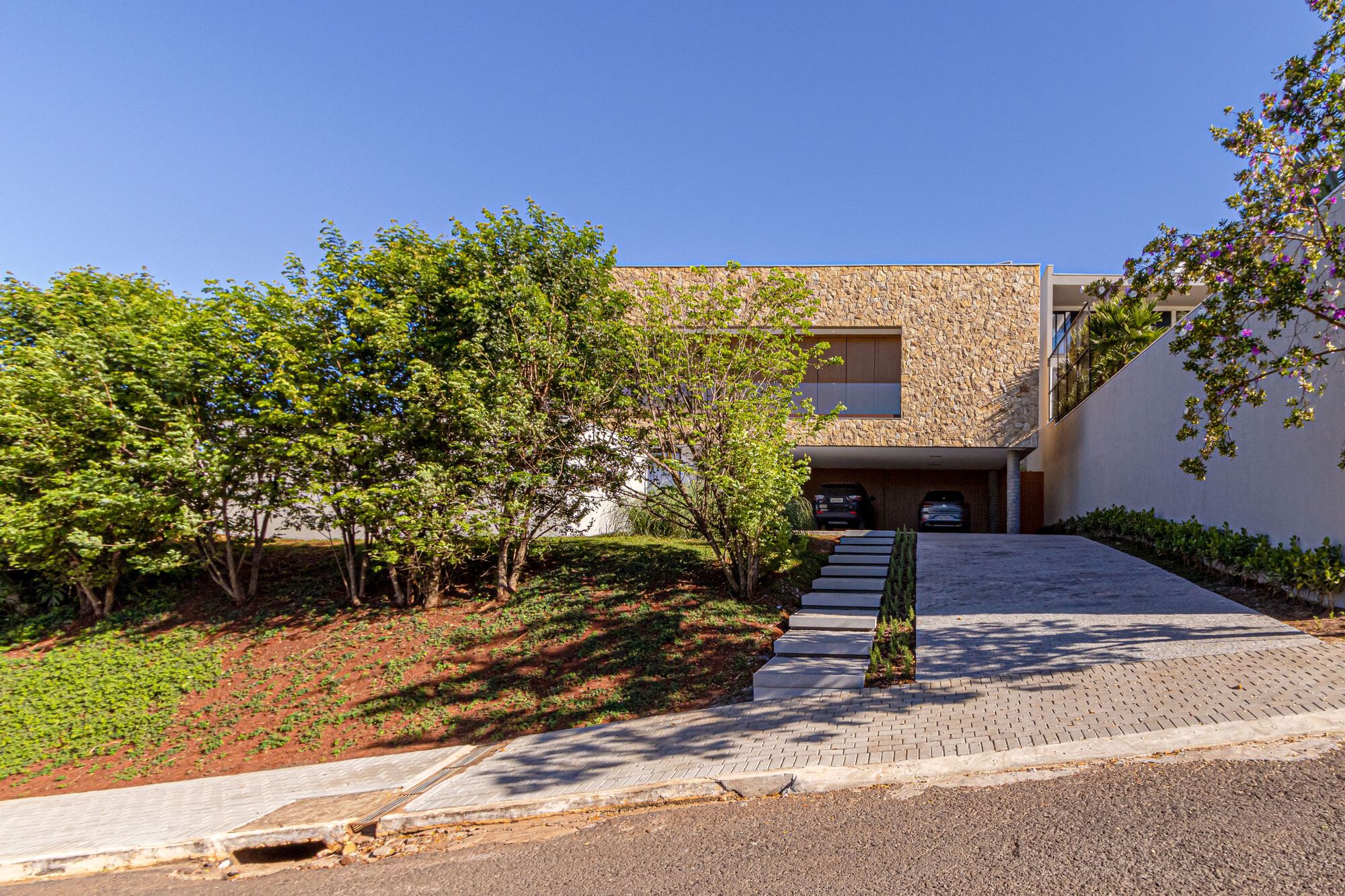 Street view of the stone-clad facade with sloped lawn and concrete steps under clear blue sky