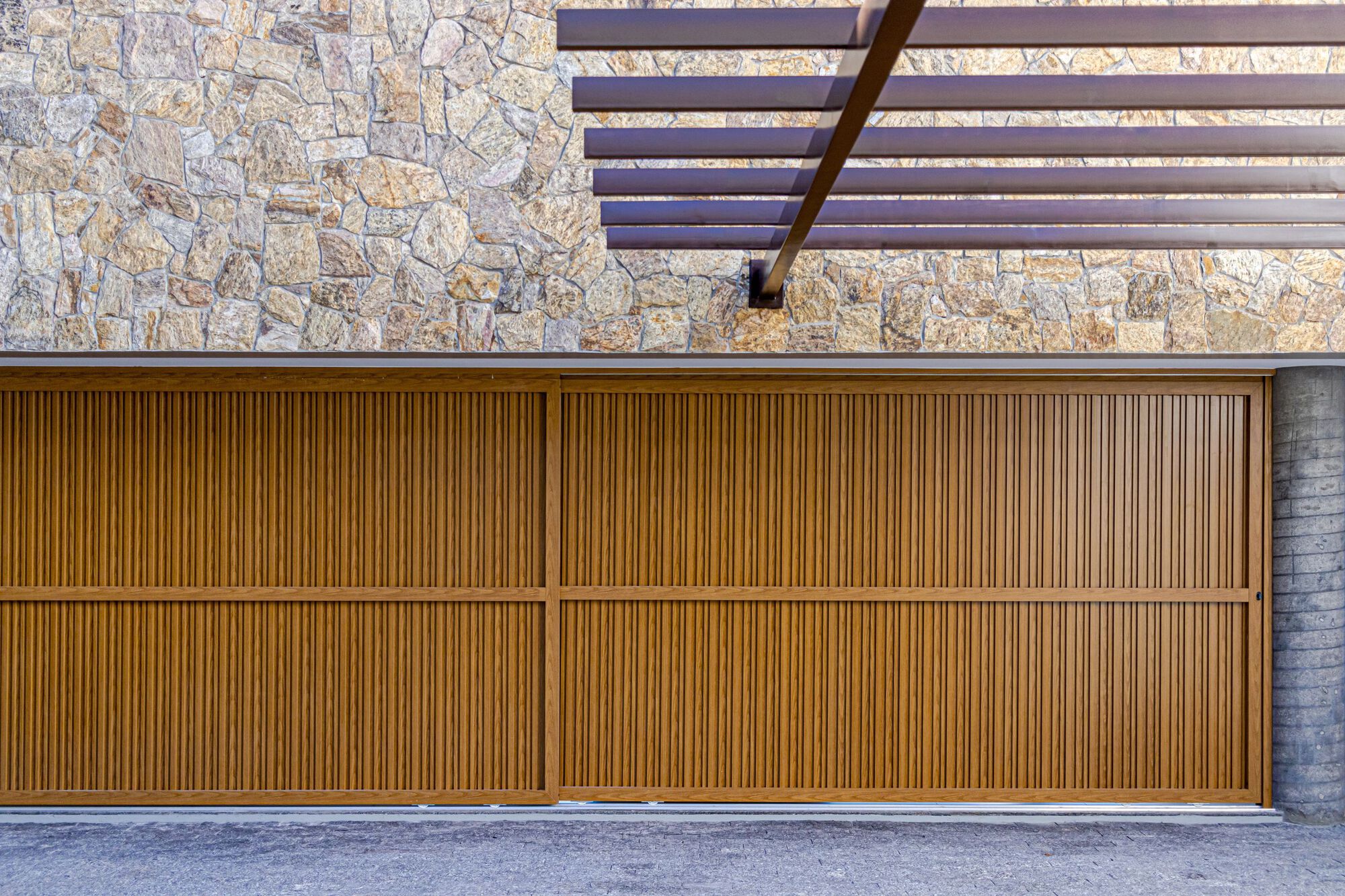 Vertical timber slat garage door beneath a slatted pergola and rough stone wall in daylight