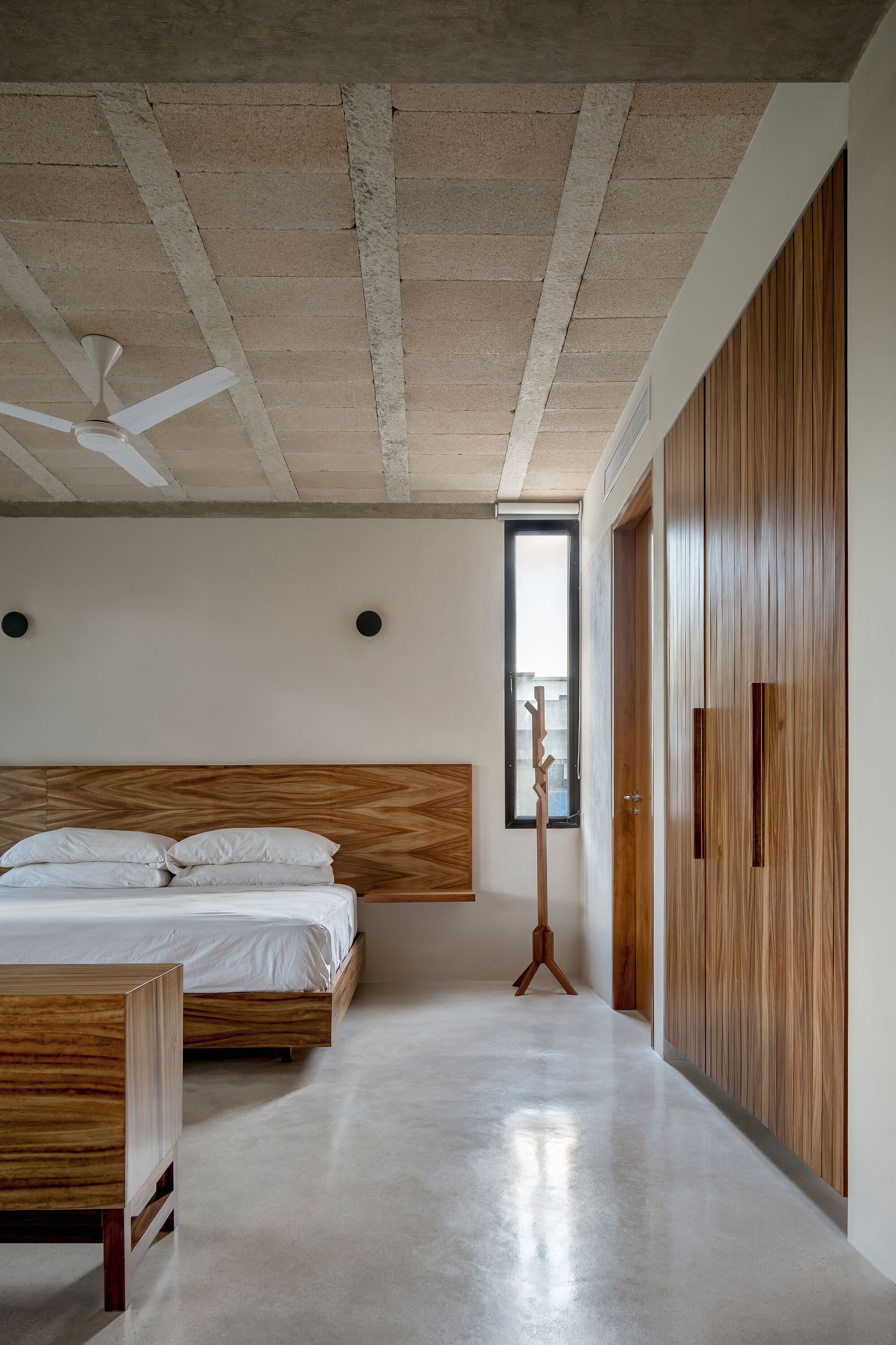 Bedroom with exposed concrete ceiling beams, vertical timber paneling and polished concrete floor in daylight