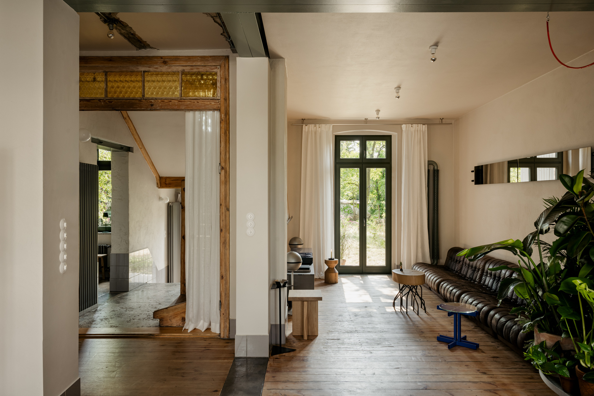 Open interior with exposed timber framing, white curtains, and tall black-framed doors leading to greenery outside
