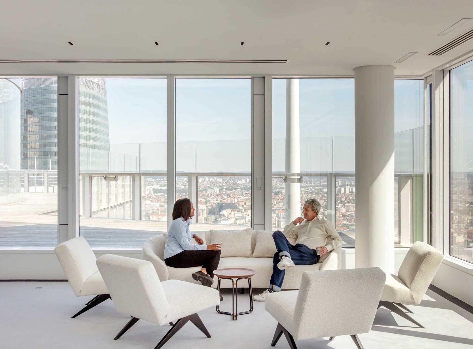 High-rise lounge with floor-to-ceiling glass, white upholstered seating and two people conversing overlooking the city