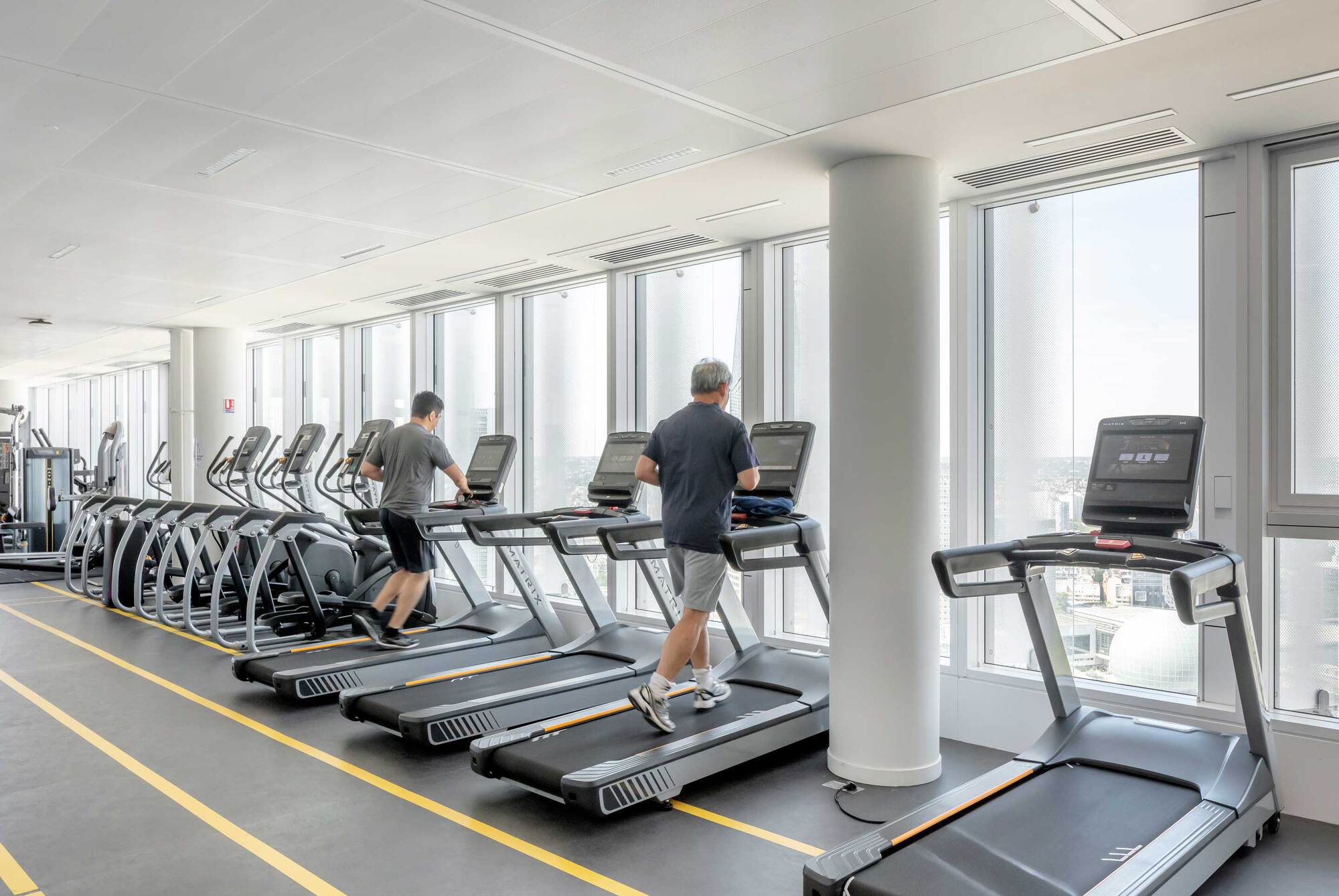 Fitness center with treadmills along the window wall and two people exercising with city views beyond