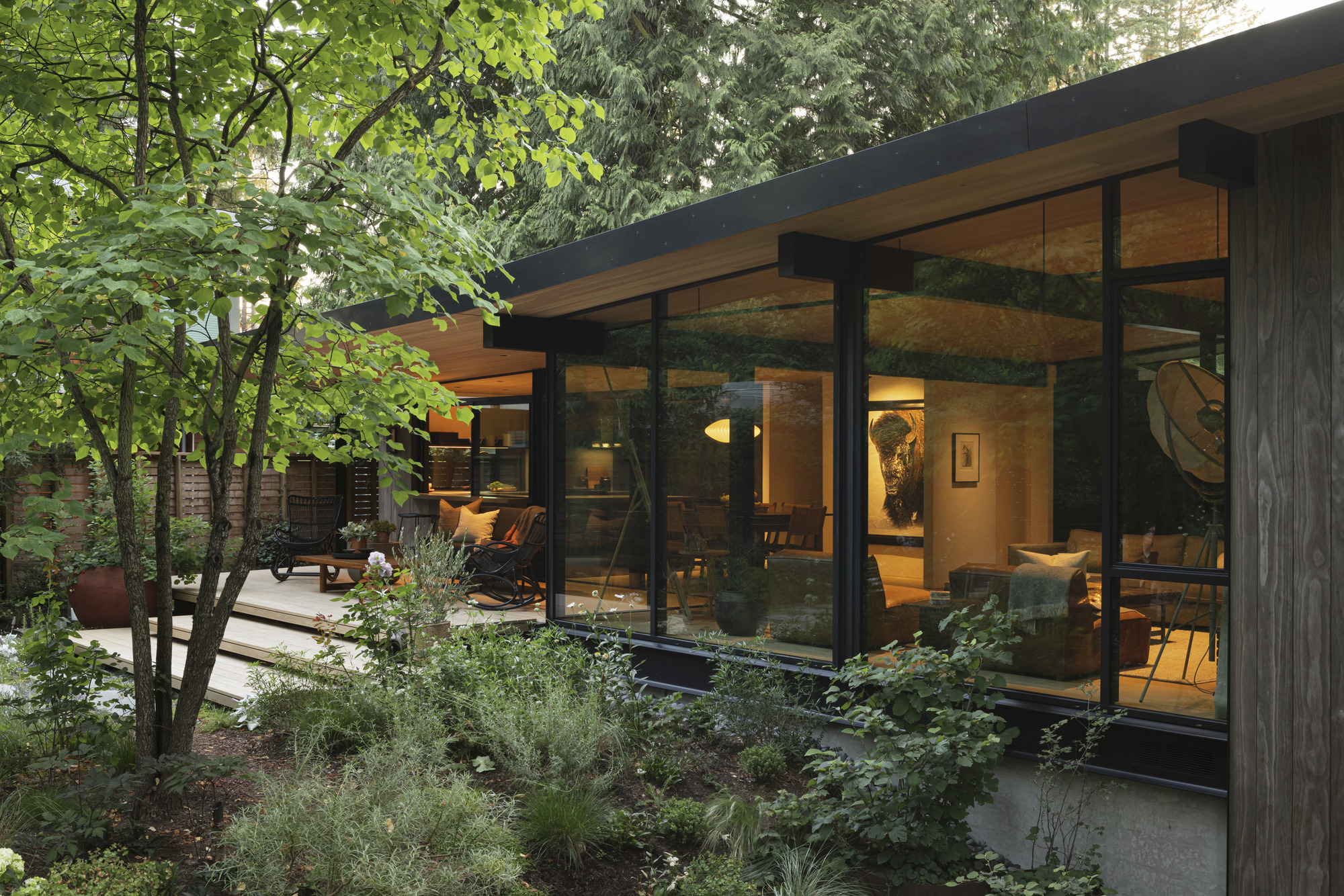 Glass-walled living pavilion framed by planted beds and deciduous trees in evening light