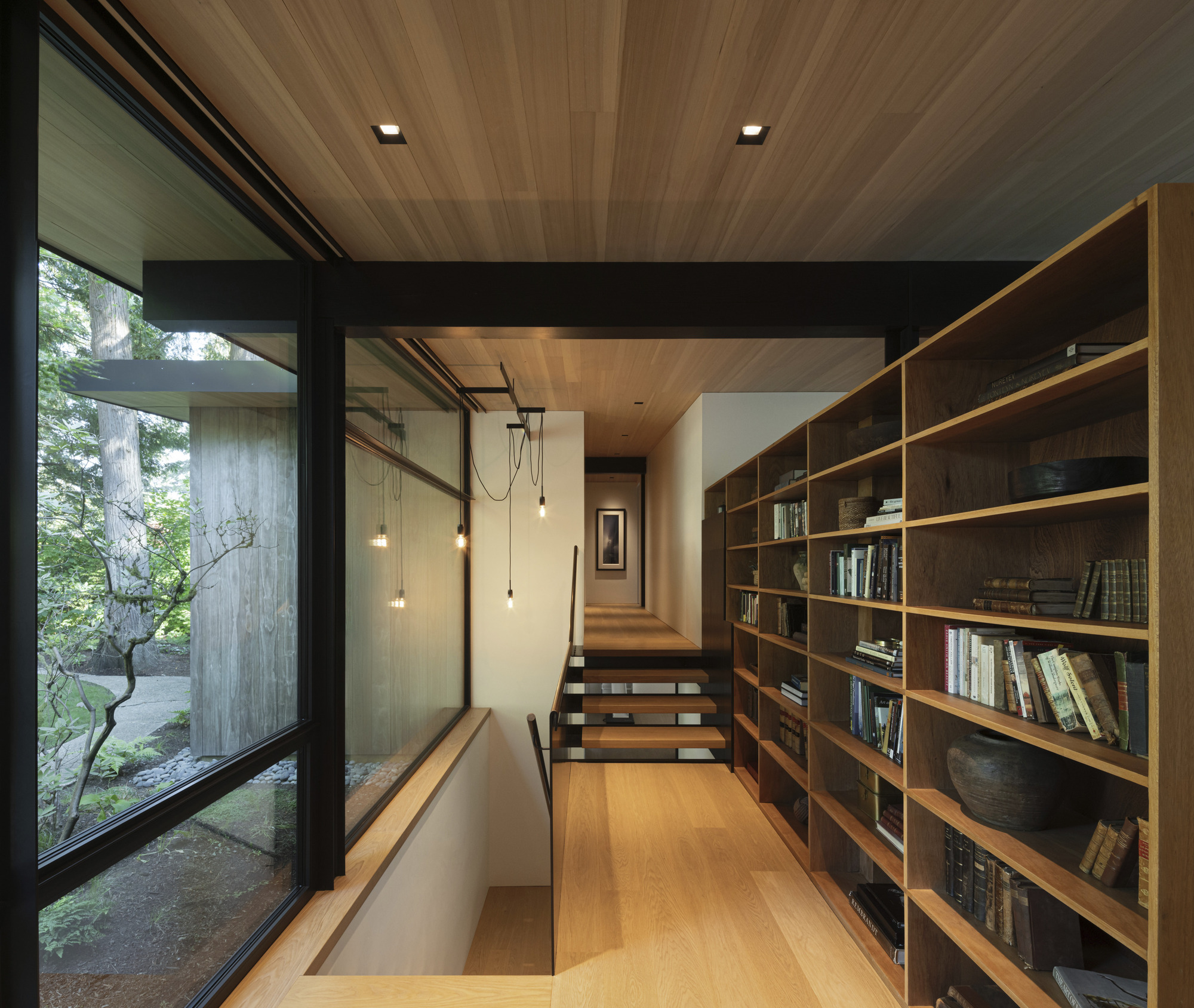 Timber-lined hallway with built-in bookshelf and pendant lights ascending to upper level