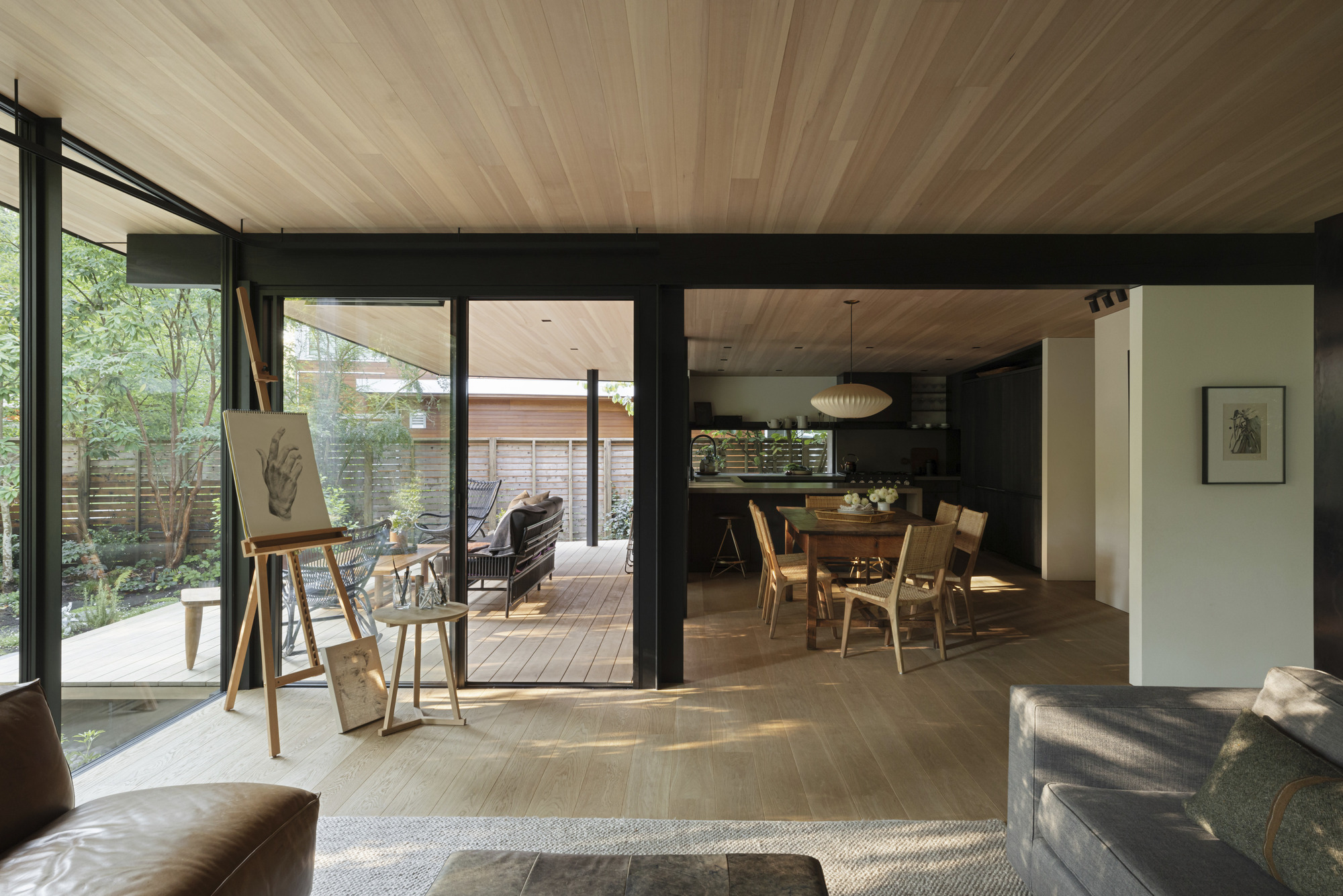 Open-plan living and dining space with black steel framing and radial wood ceiling under dappled sunlight