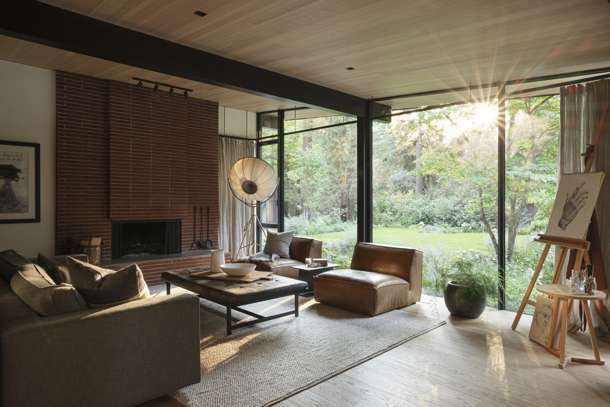 Living room with brick fireplace and corner windows framing views of trees in afternoon light