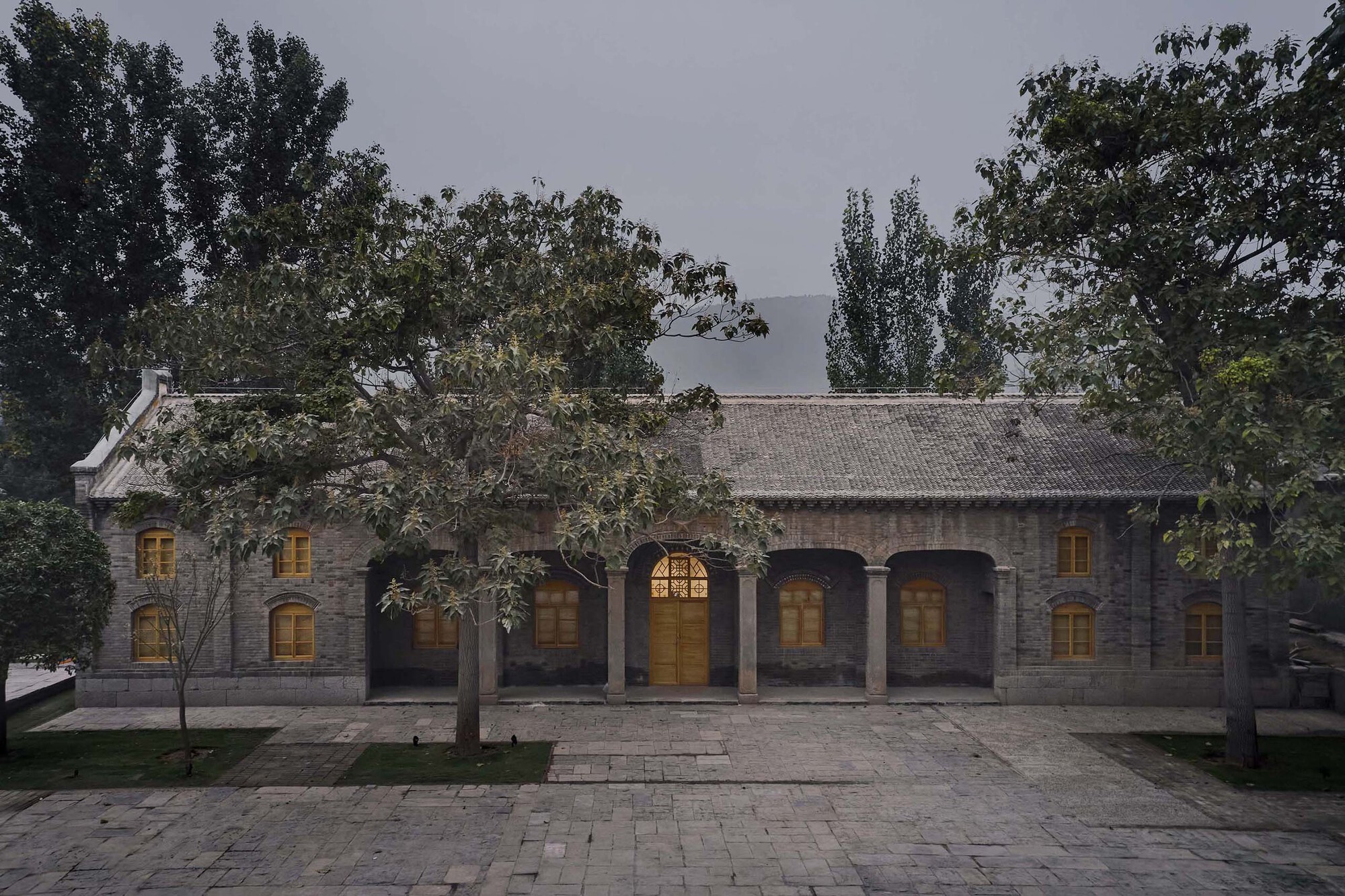 Restored brick facade with arched openings and tiled roof illuminated at dusk beneath overhanging trees
