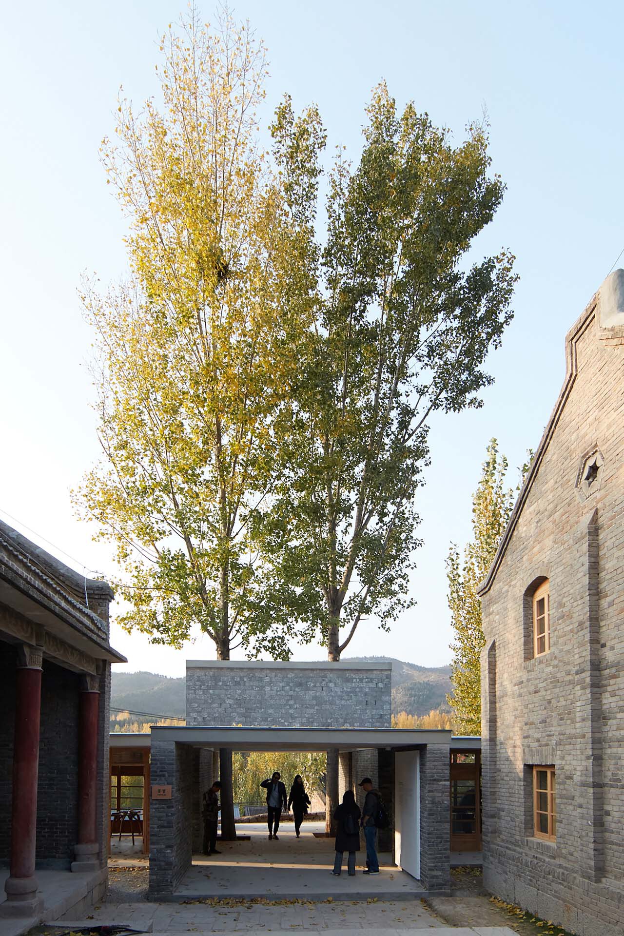 Grey brick pavilion with flat roof connecting two existing structures under tall trees in autumn