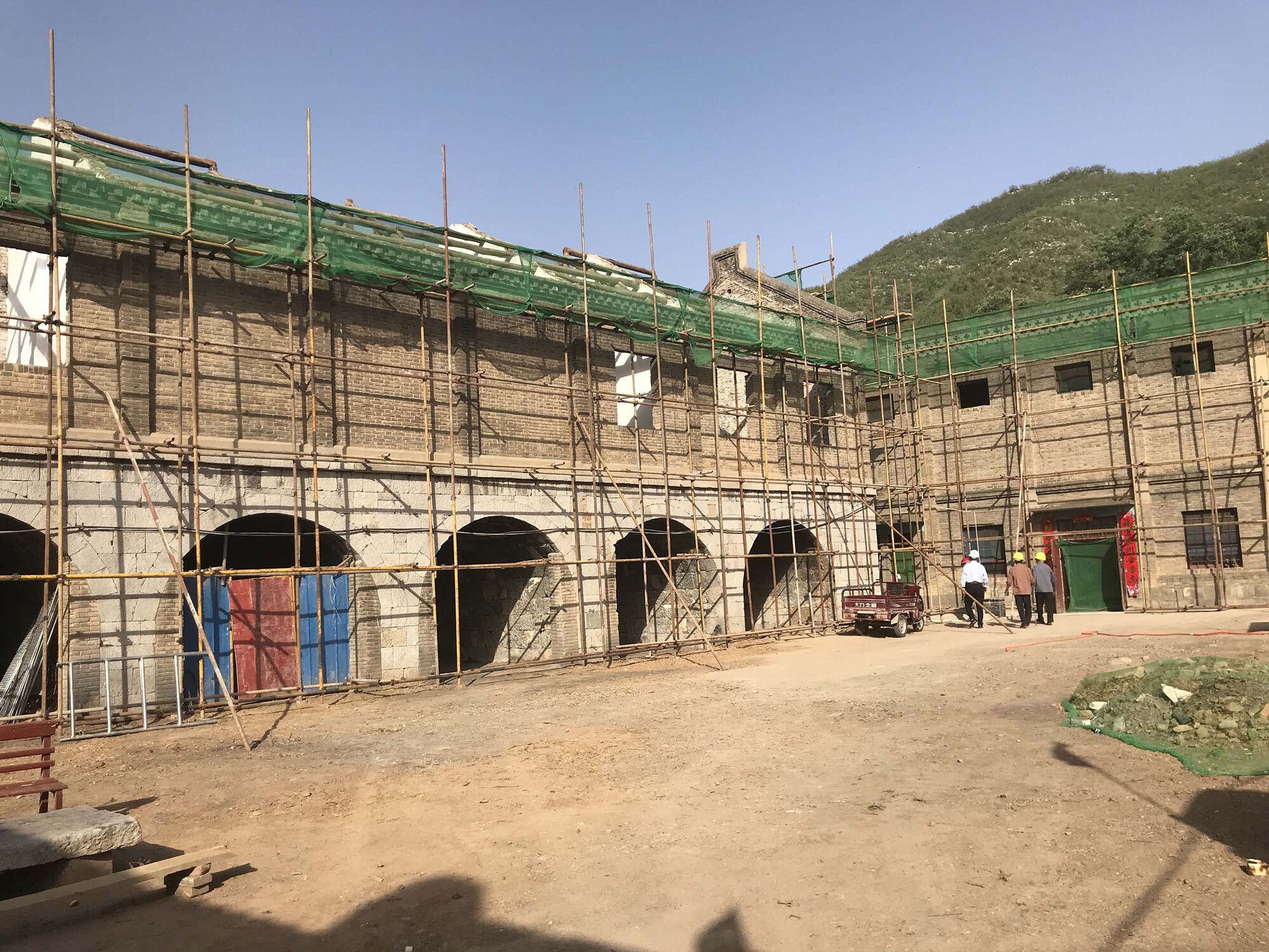 Renovation site showing arched openings wrapped in bamboo scaffolding and green mesh netting with hillside beyond