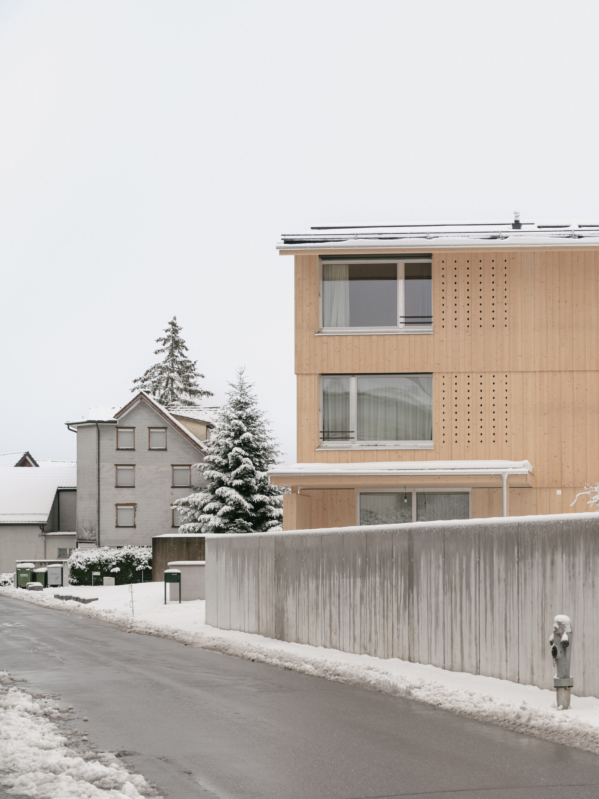 Timber facade with perforated screens beside horizontal windows backed by a weathered concrete boundary wall