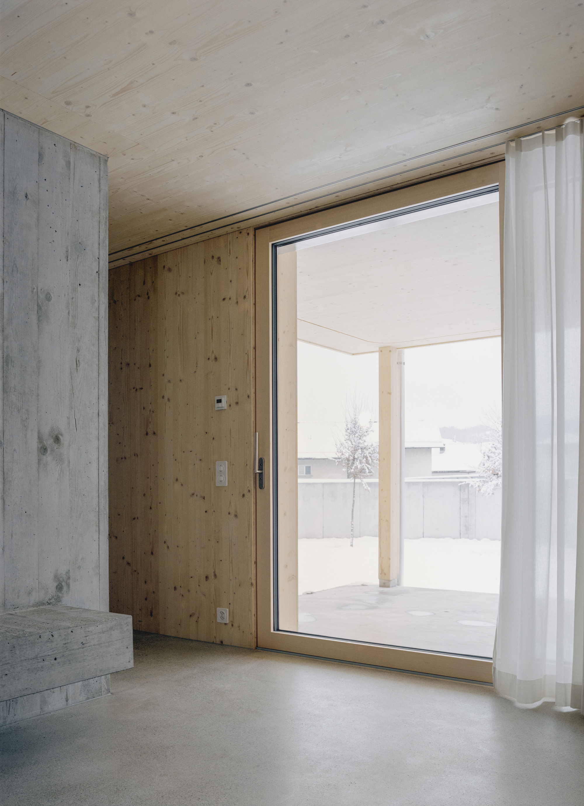 Interior room with plywood ceiling and concrete walls framing a glazed door to a snow-dusted terrace