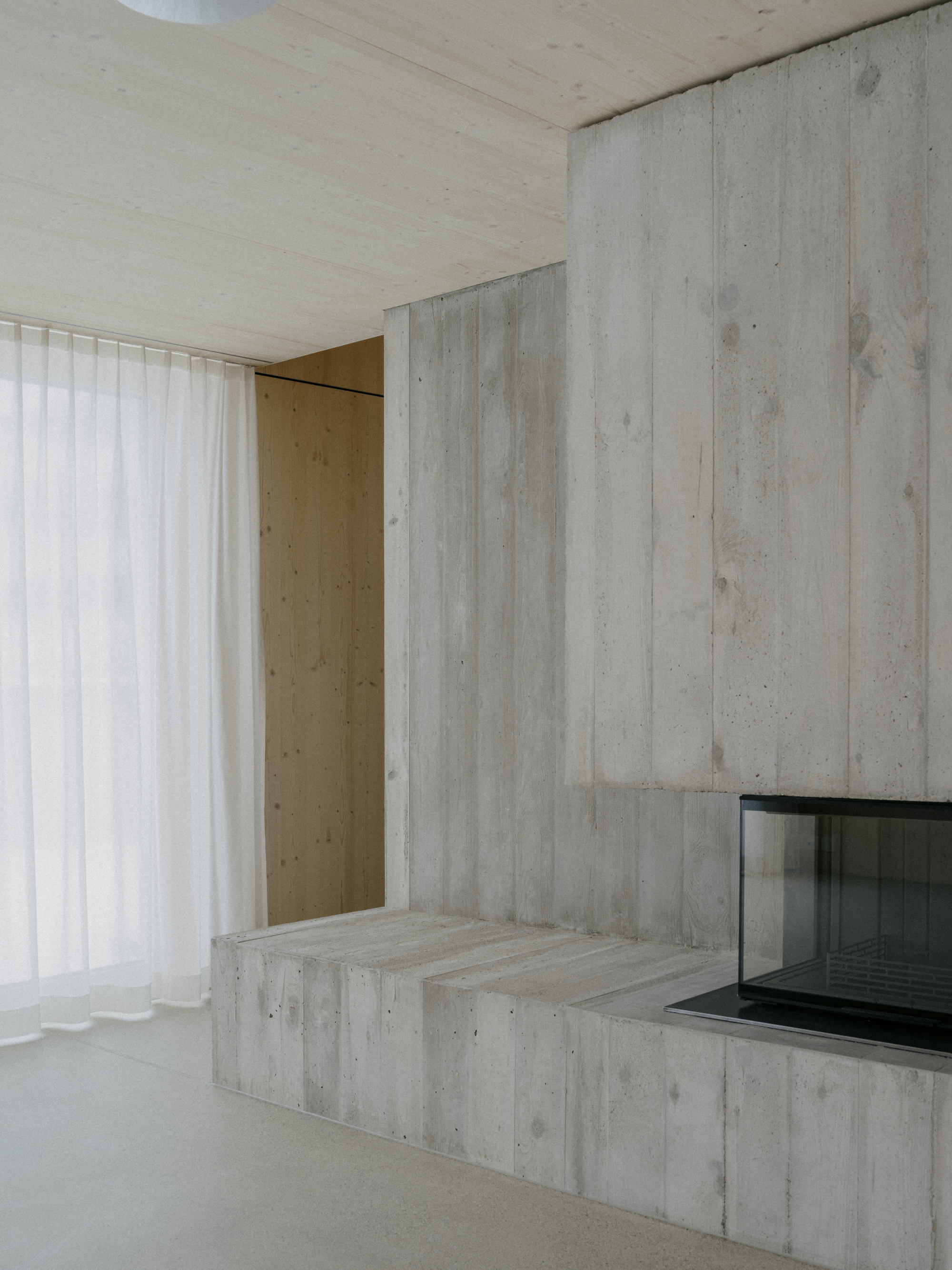 Board-formed concrete fireplace bench and wall beside a translucent curtain and plywood ceiling