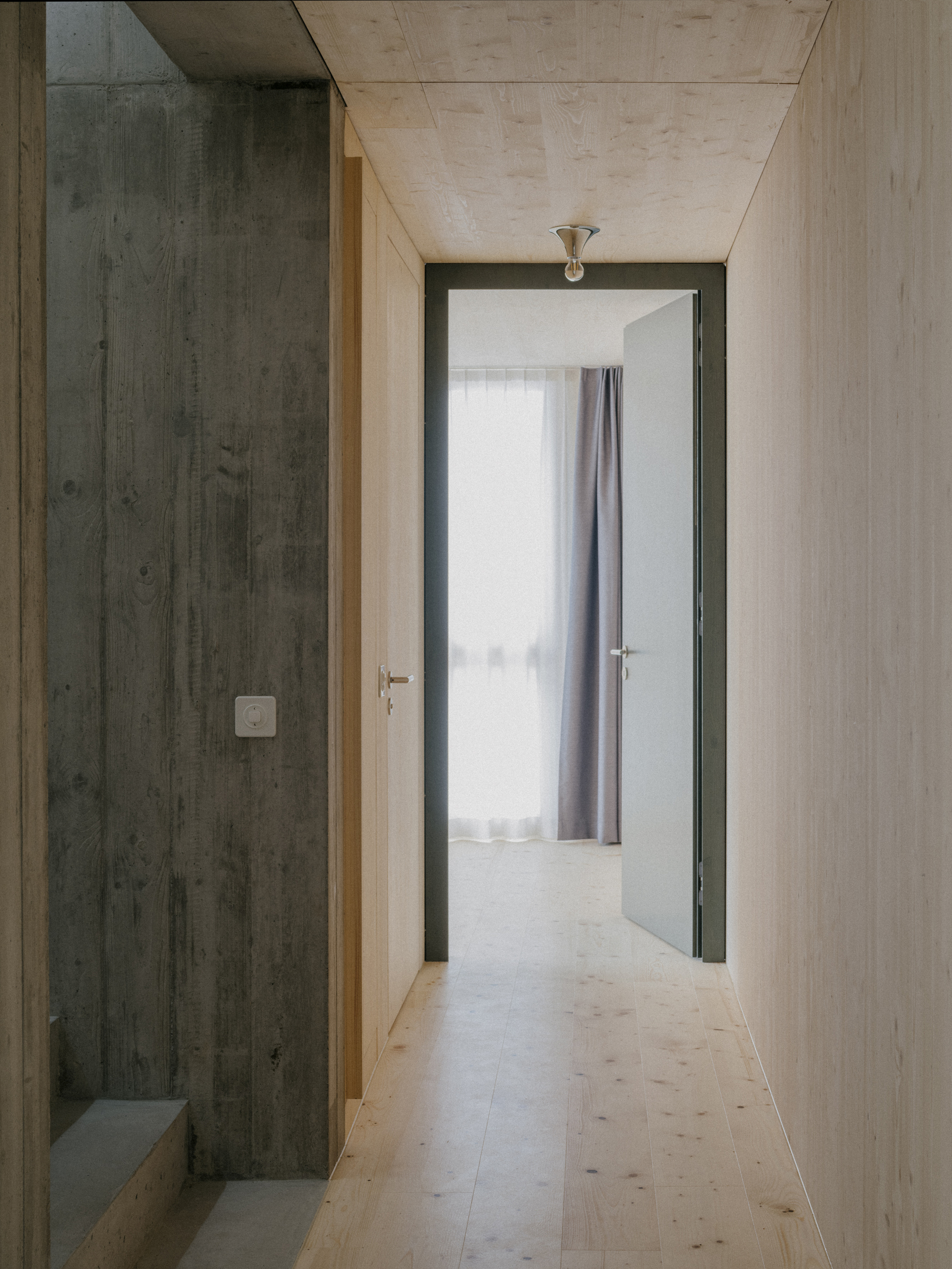 Narrow hallway with light plywood and dark timber walls leading to a room with sheer curtains