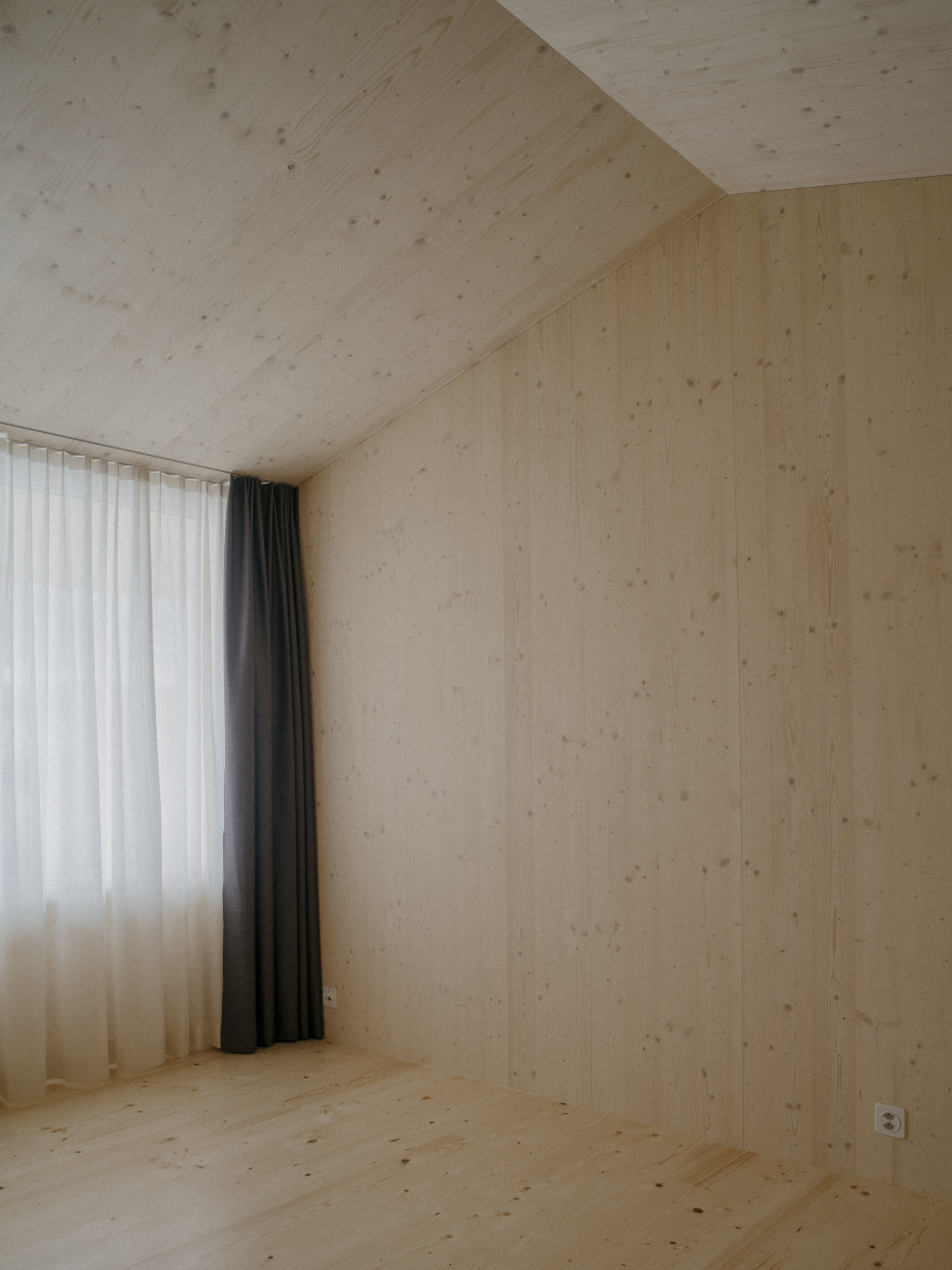 Interior corner of pale timber-lined room with sloped ceiling and layered white and grey curtains