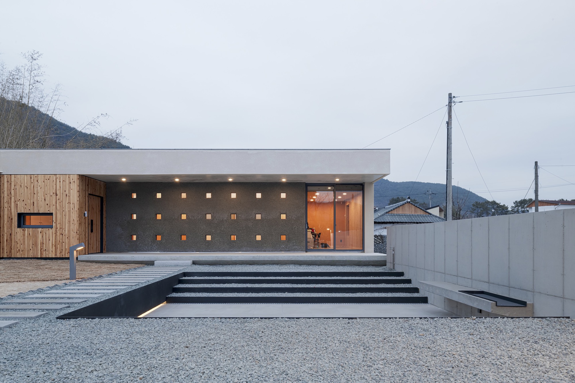 Entry facade with perforated concrete wall and wide stairs at dusk under overcast sky