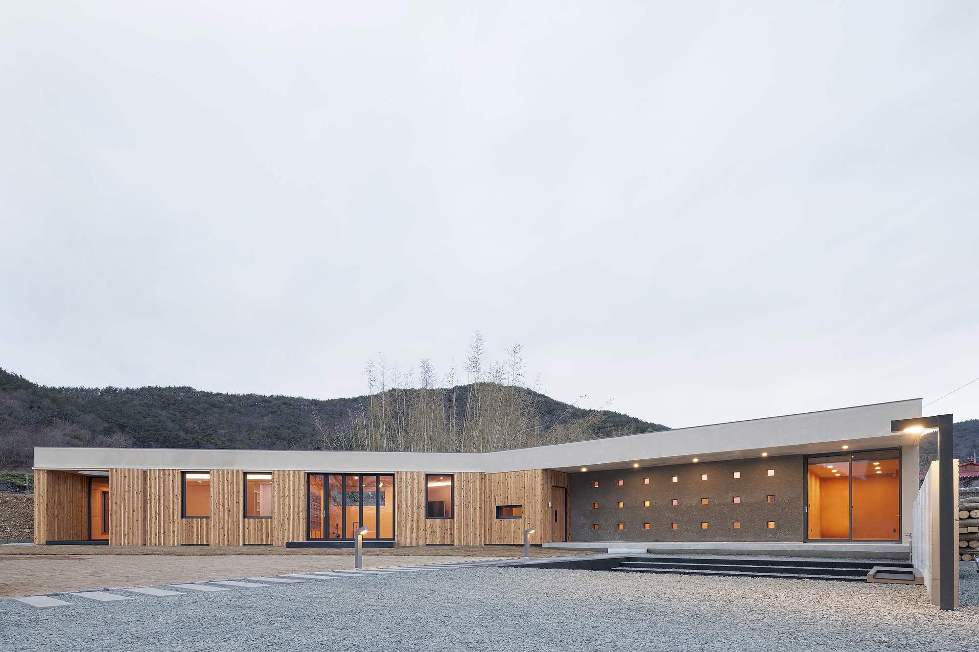 Horizontal timber-clad facade with scattered punched windows and illuminated concrete porch at dusk