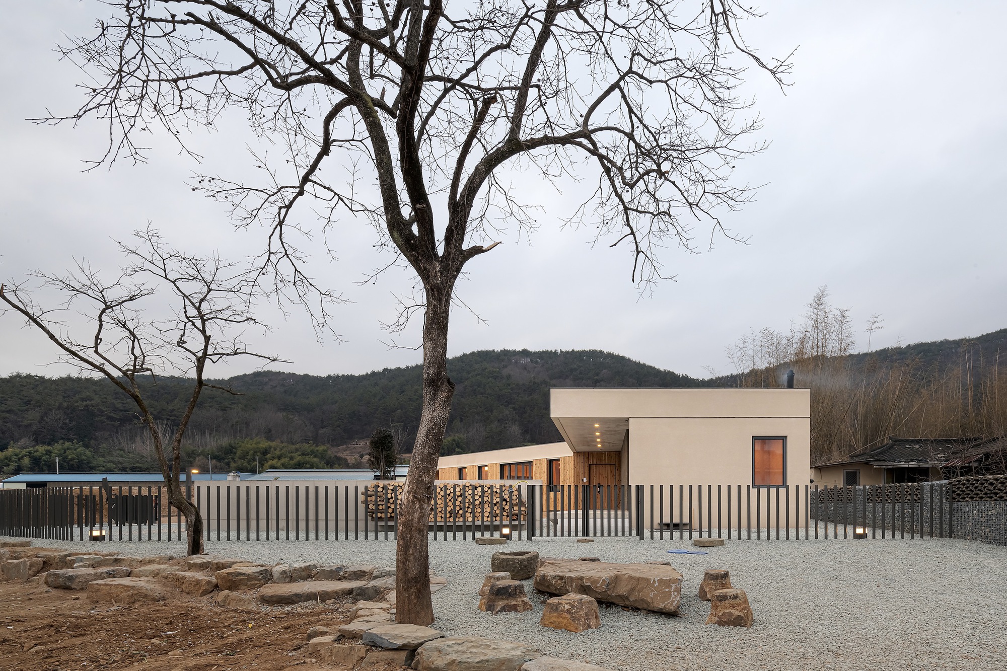 White concrete house behind vertical slatted fence with bare winter tree and hillside backdrop