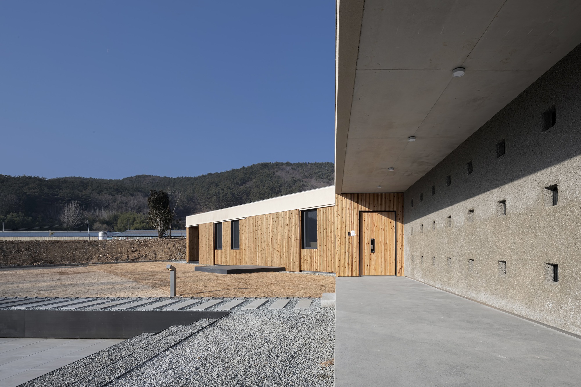 Covered walkway with concrete soffit beside gravel court and timber-clad wing under clear sky