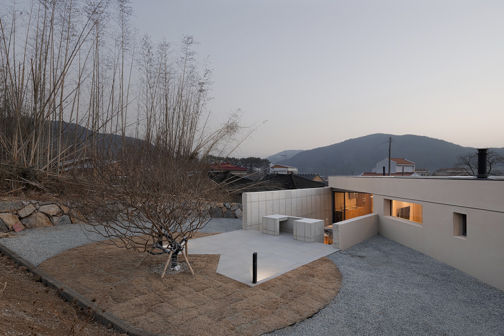 Courtyard view with gravel ground, bare winter tree and white concrete seating under a dusk sky
