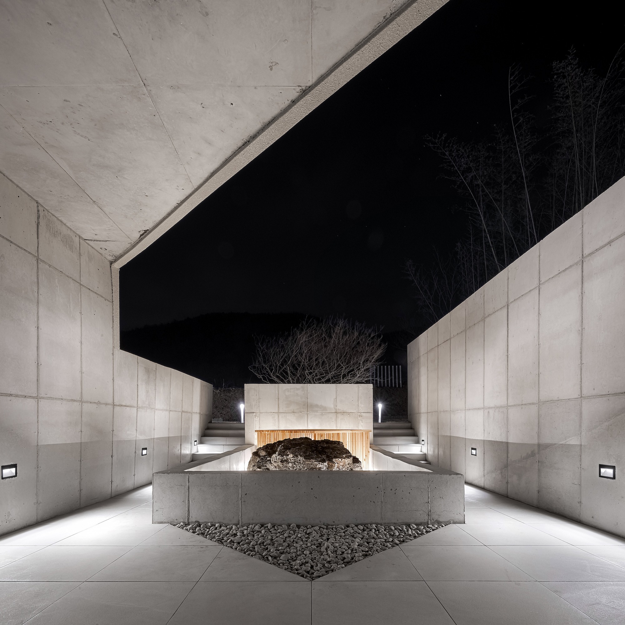 Covered entrance passage with concrete walls, overhead beams and a central planted tree at night