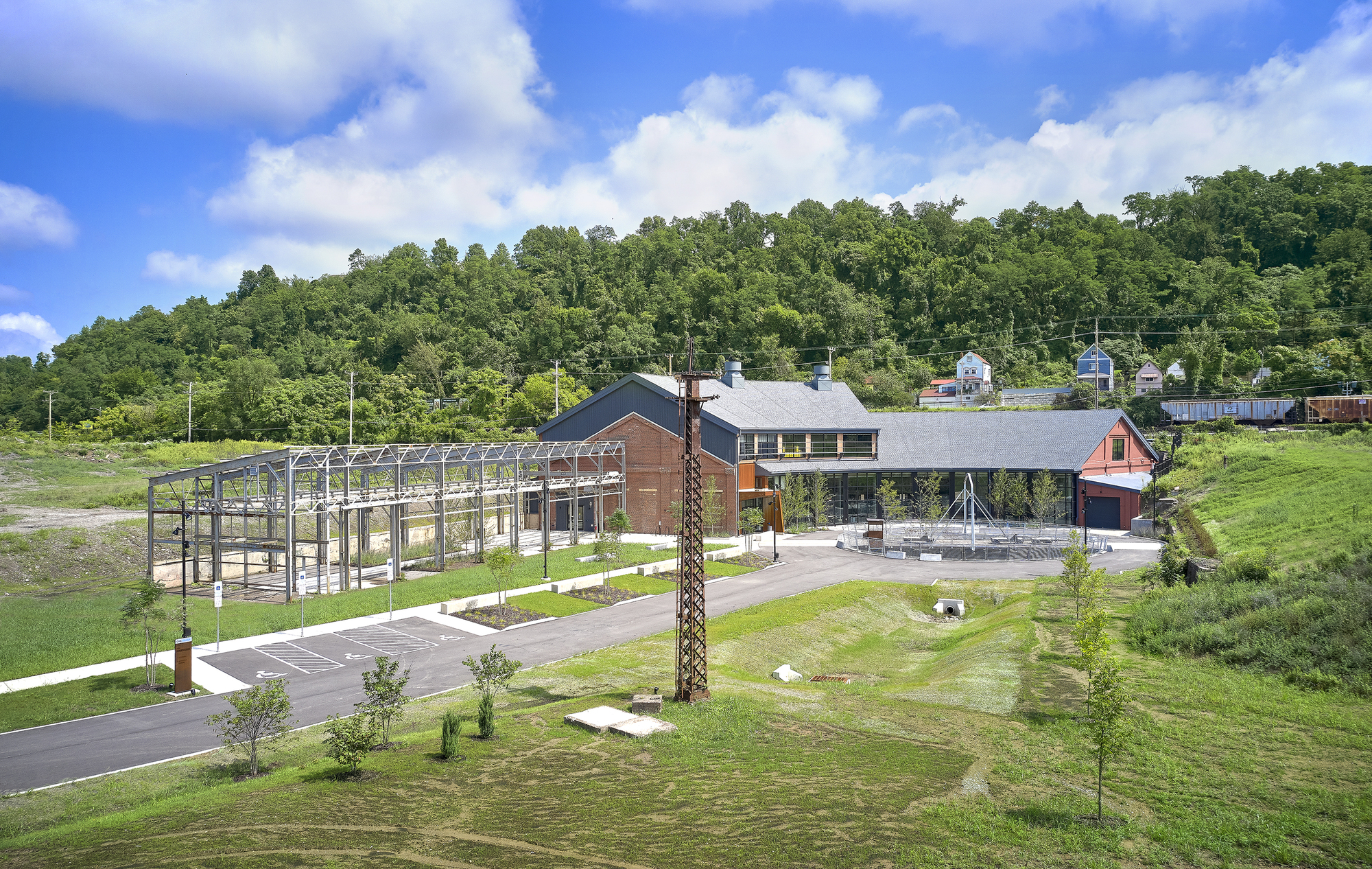 Site view showing the main building with weathered steel panels, glass facades and an open metal frame structure on the left