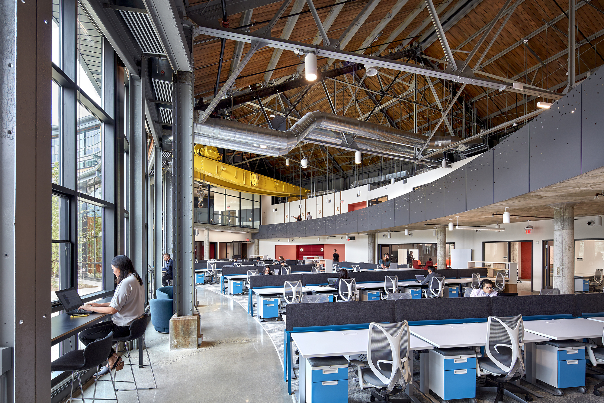 Double-height workspace with exposed wood bowstring trusses, concrete columns and rows of open office workstations below a mezzanine