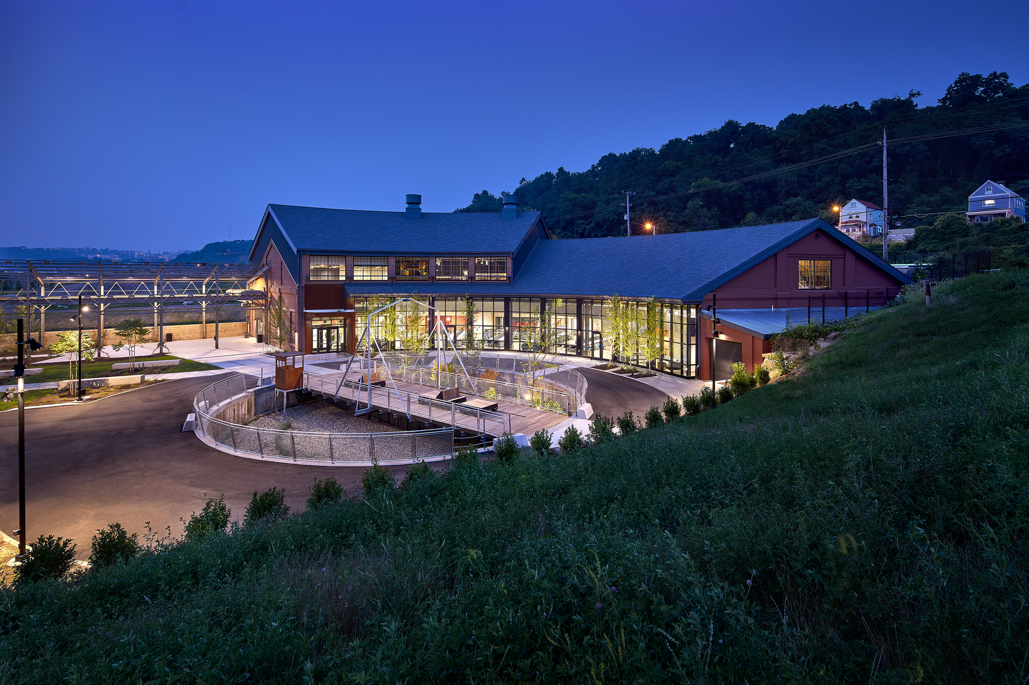 Glass-walled entrance with timber-framed gabled roofs and circular drive at dusk with hillside beyond
