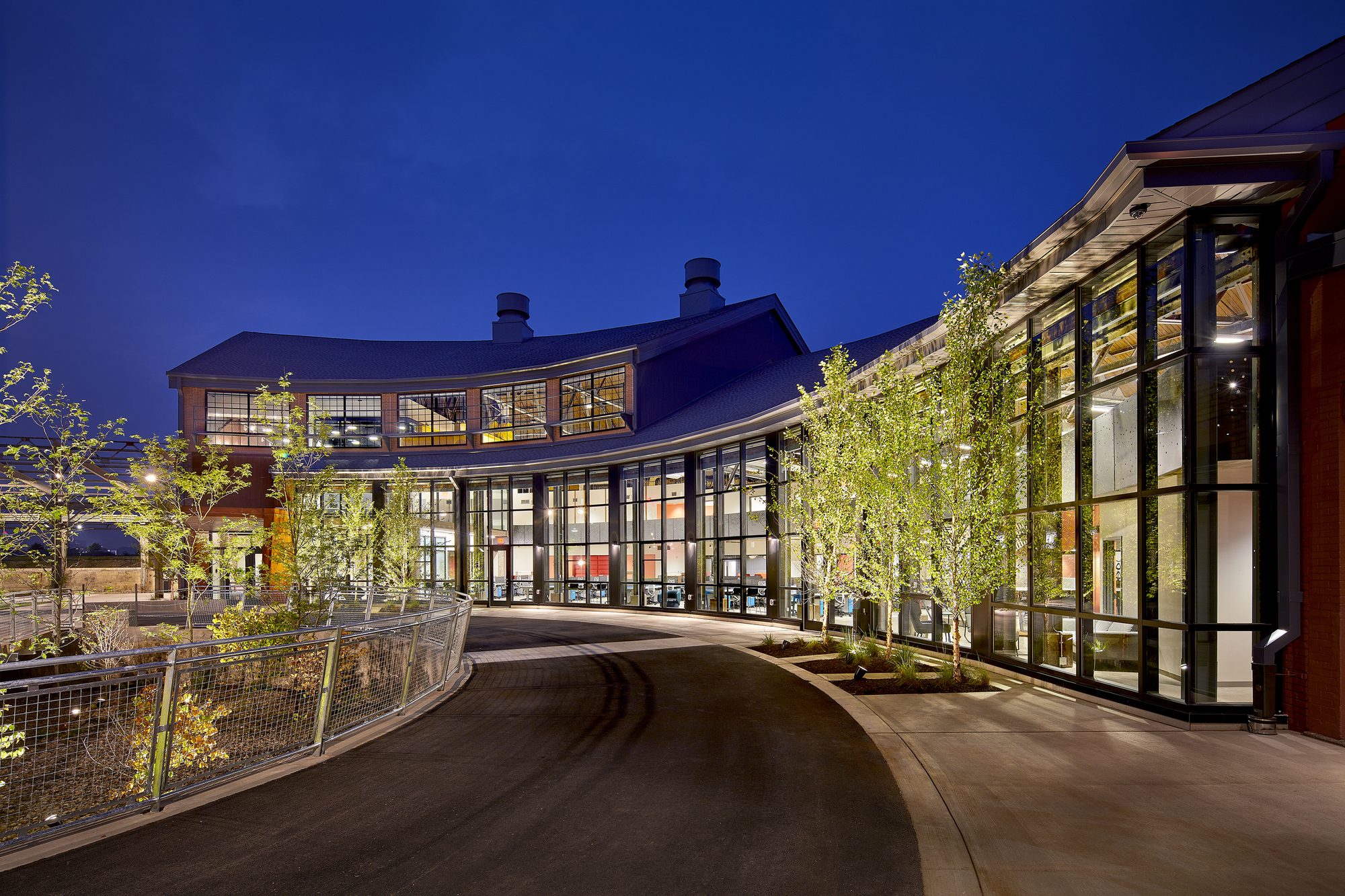 Curving entry drive with illuminated glass facades and planted courtyard under evening sky