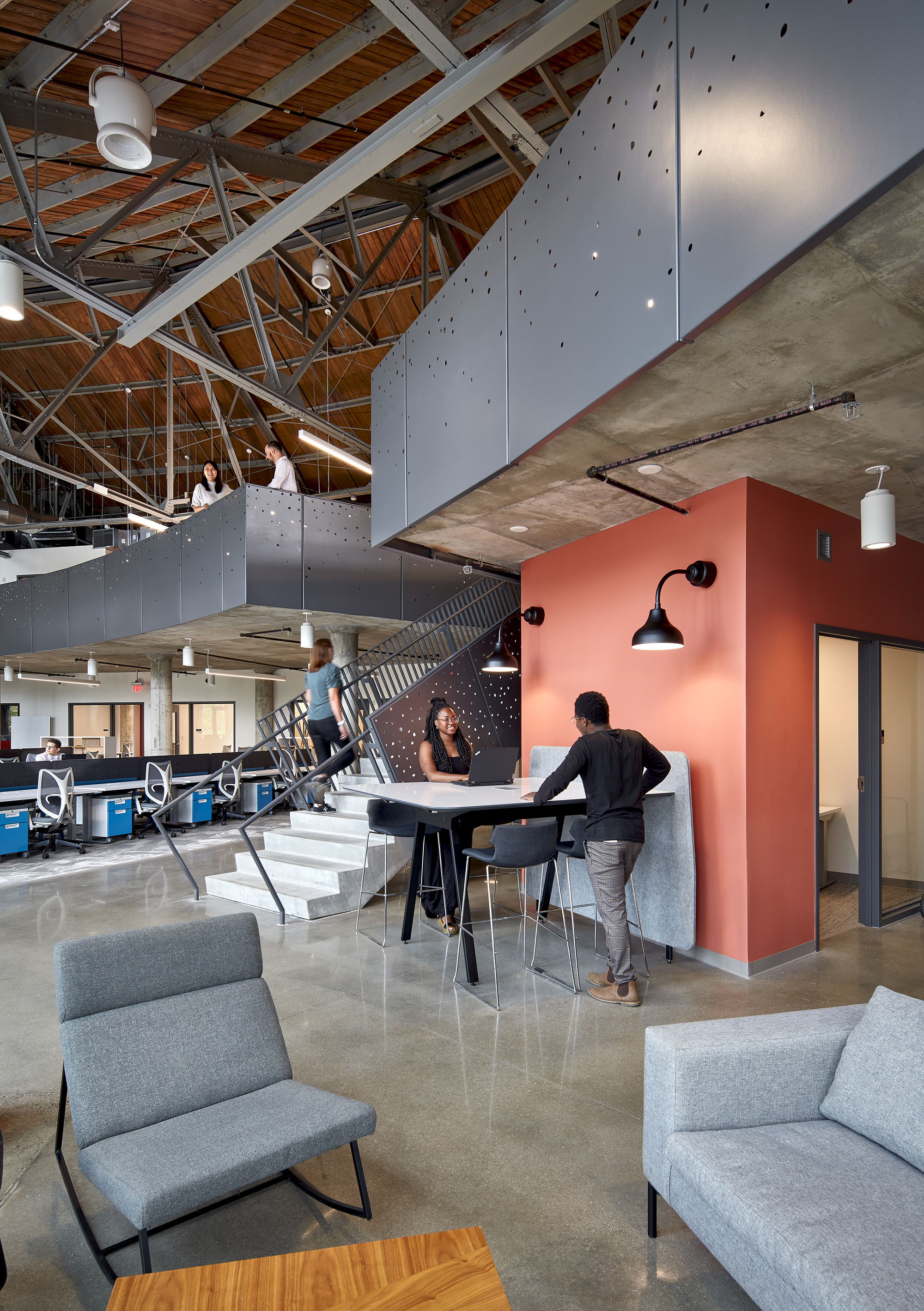Open workspace with coral-colored partitions beneath white steel trusses and timber ceiling with three people conversing