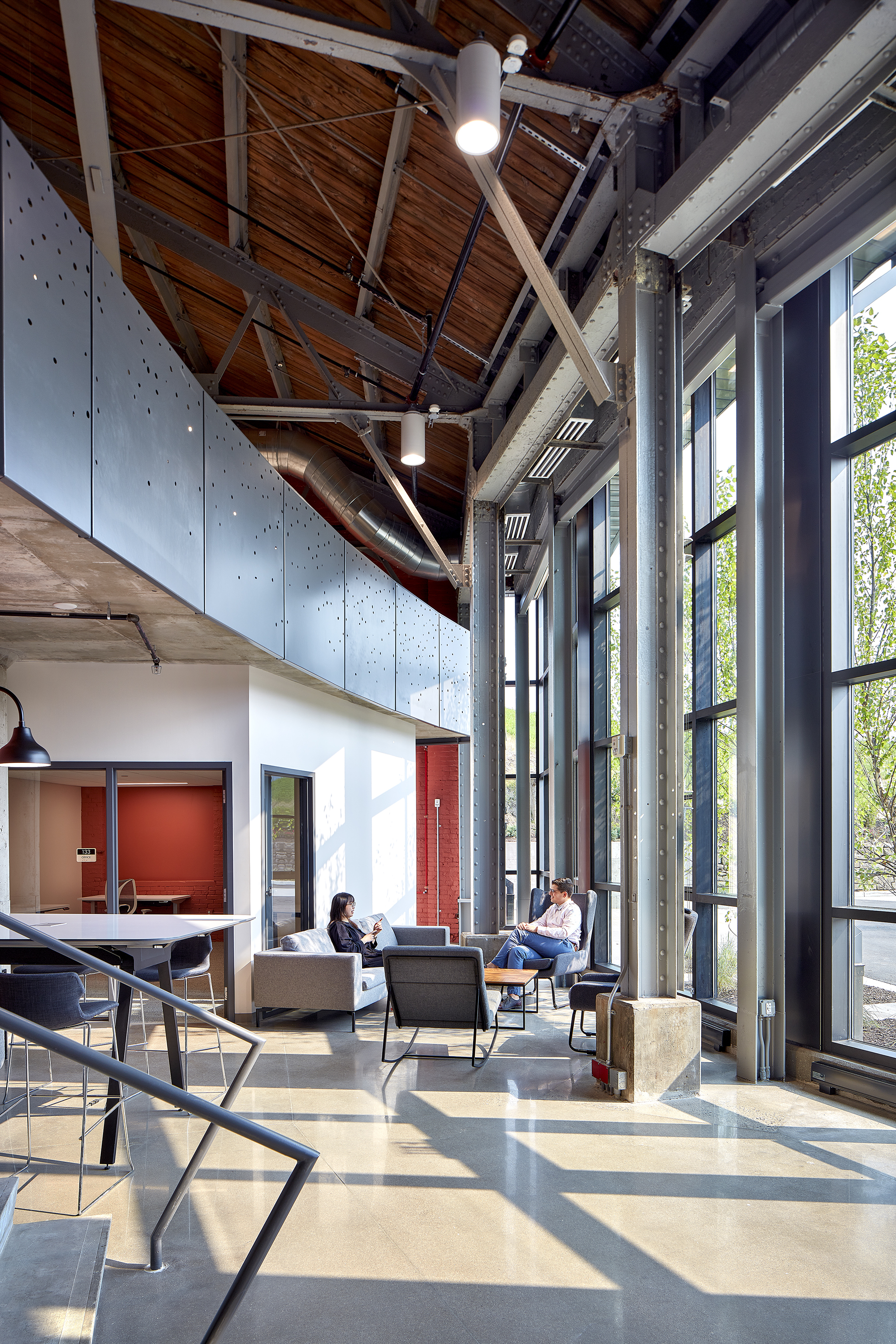 Lounge seating area beneath exposed timber trusses and metal ductwork with afternoon sunlight streaming through tall windows