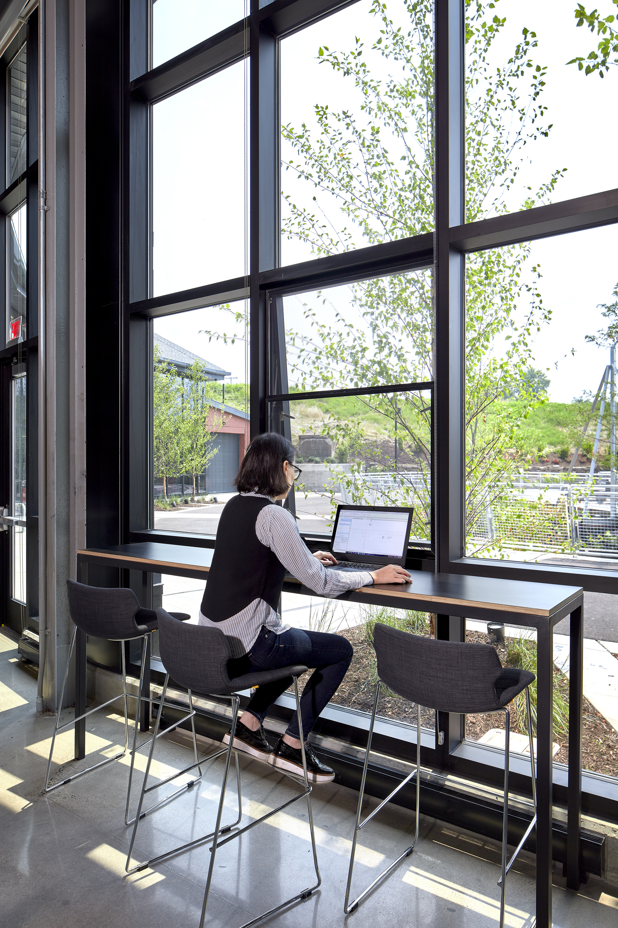 Person working at a standing-height table beside floor-to-ceiling windows overlooking exterior trees and parking lot