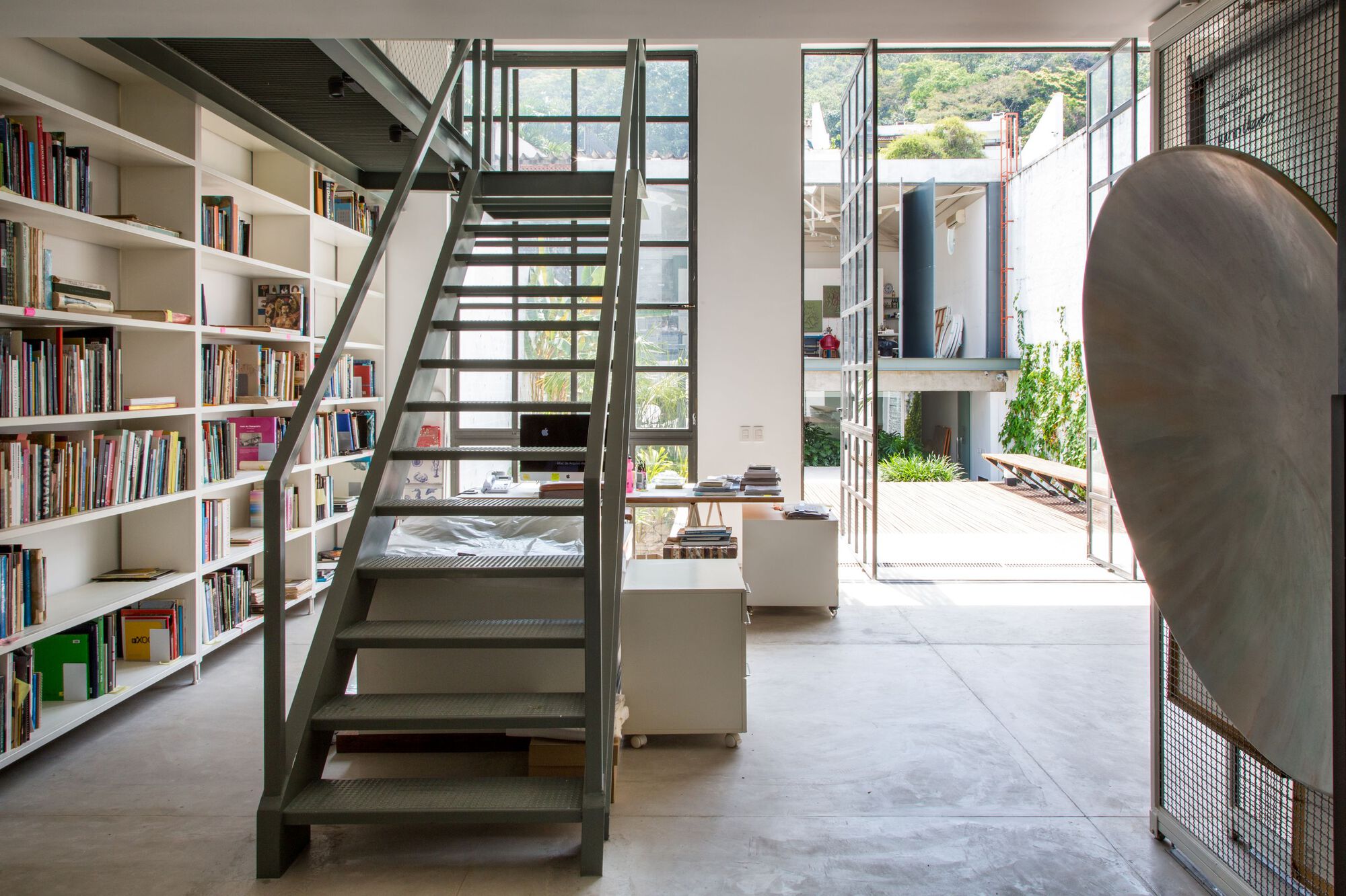 Steel staircase with perforated treads rising beside a floor-to-ceiling bookshelf in a double-height living space