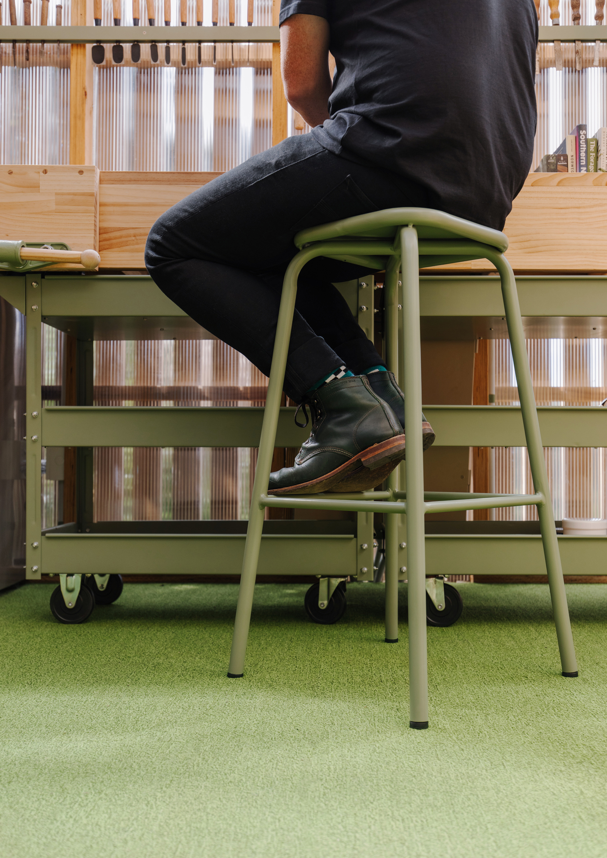 Person seated on green tubular steel stool beside rolling storage cart on artificial turf flooring