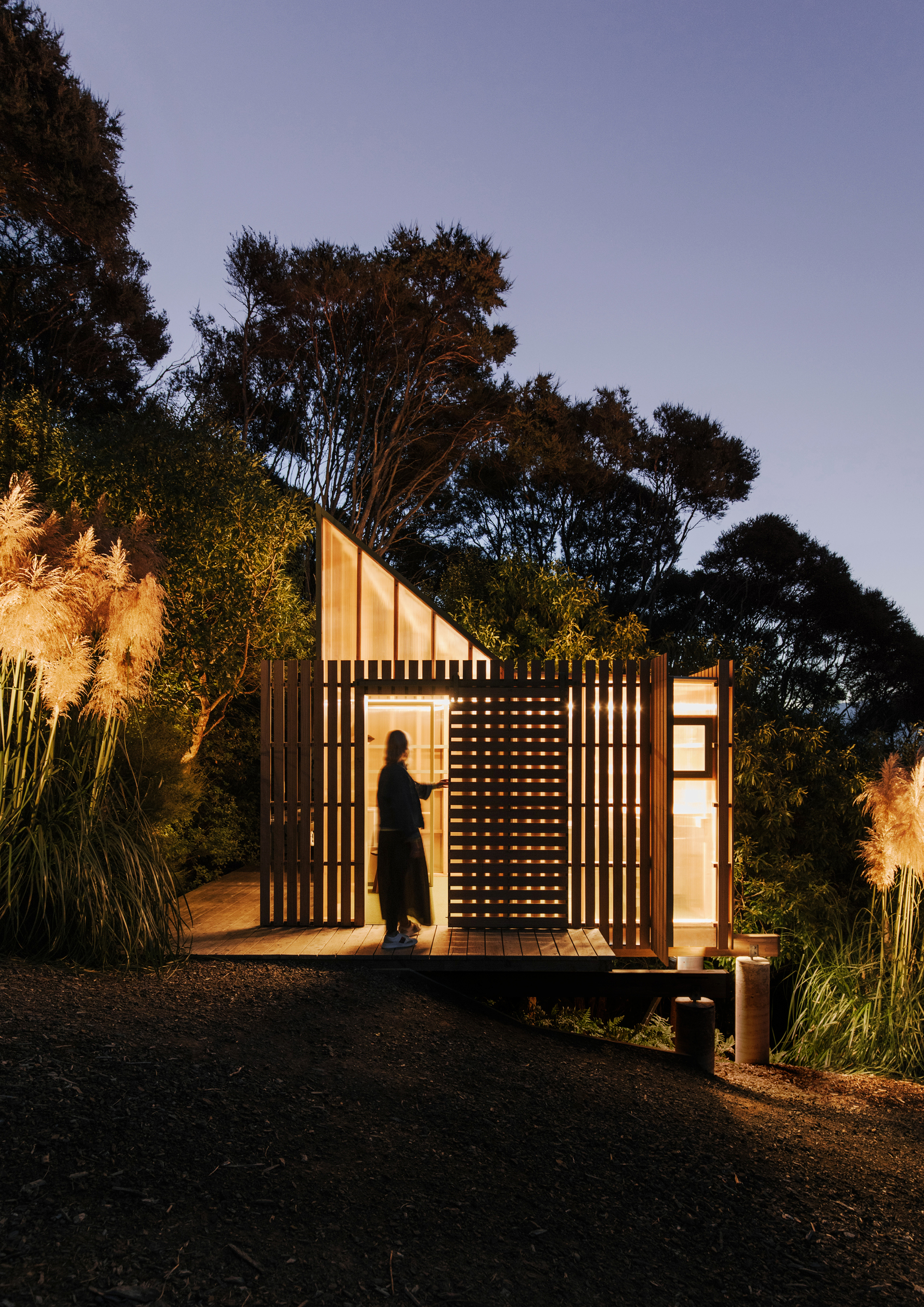 Small cabin with vertical timber screens and translucent panels glowing at dusk among native plantings
