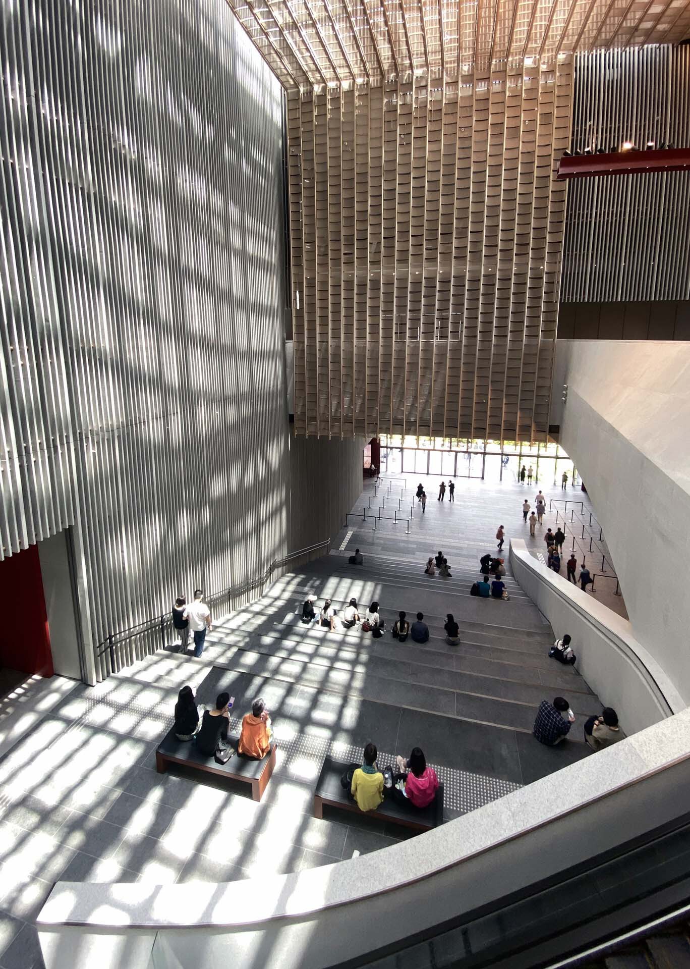 Multi-story atrium with vertical fin screens casting shadow patterns on visitors seated on steps