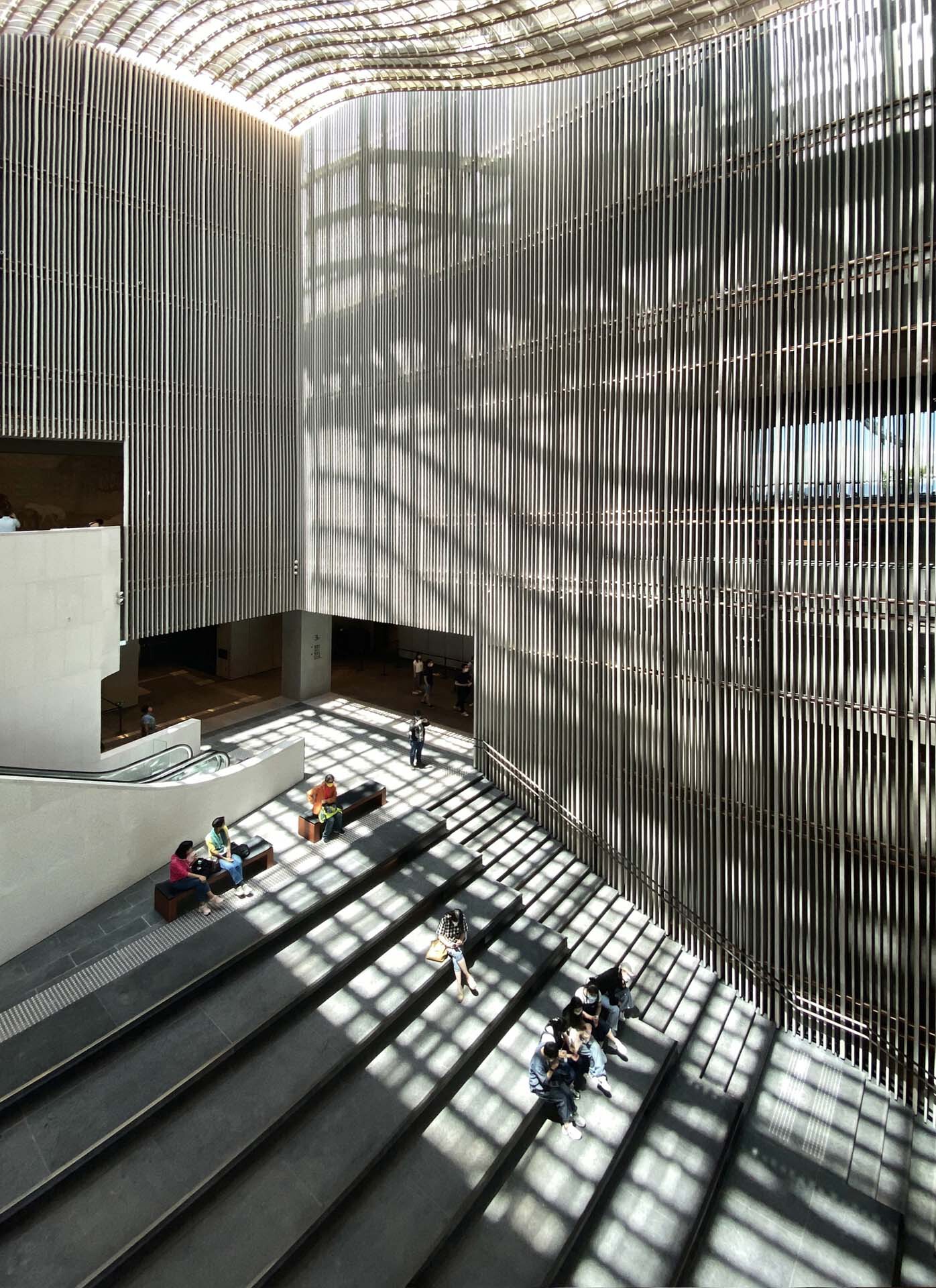 Double-height atrium with vertical metal louvers casting grid shadows on the stone floor with visitors below