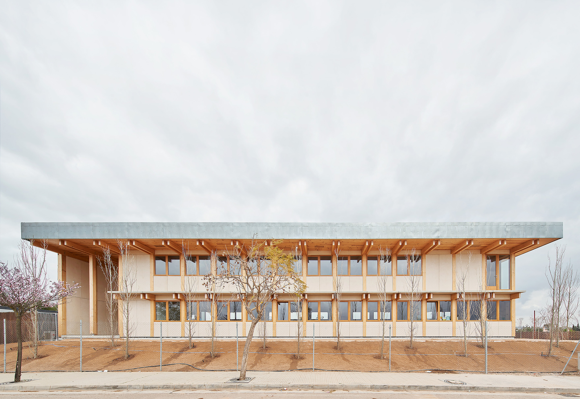 Two-story timber-framed facade with deep overhanging roof and newly planted trees in bare earth