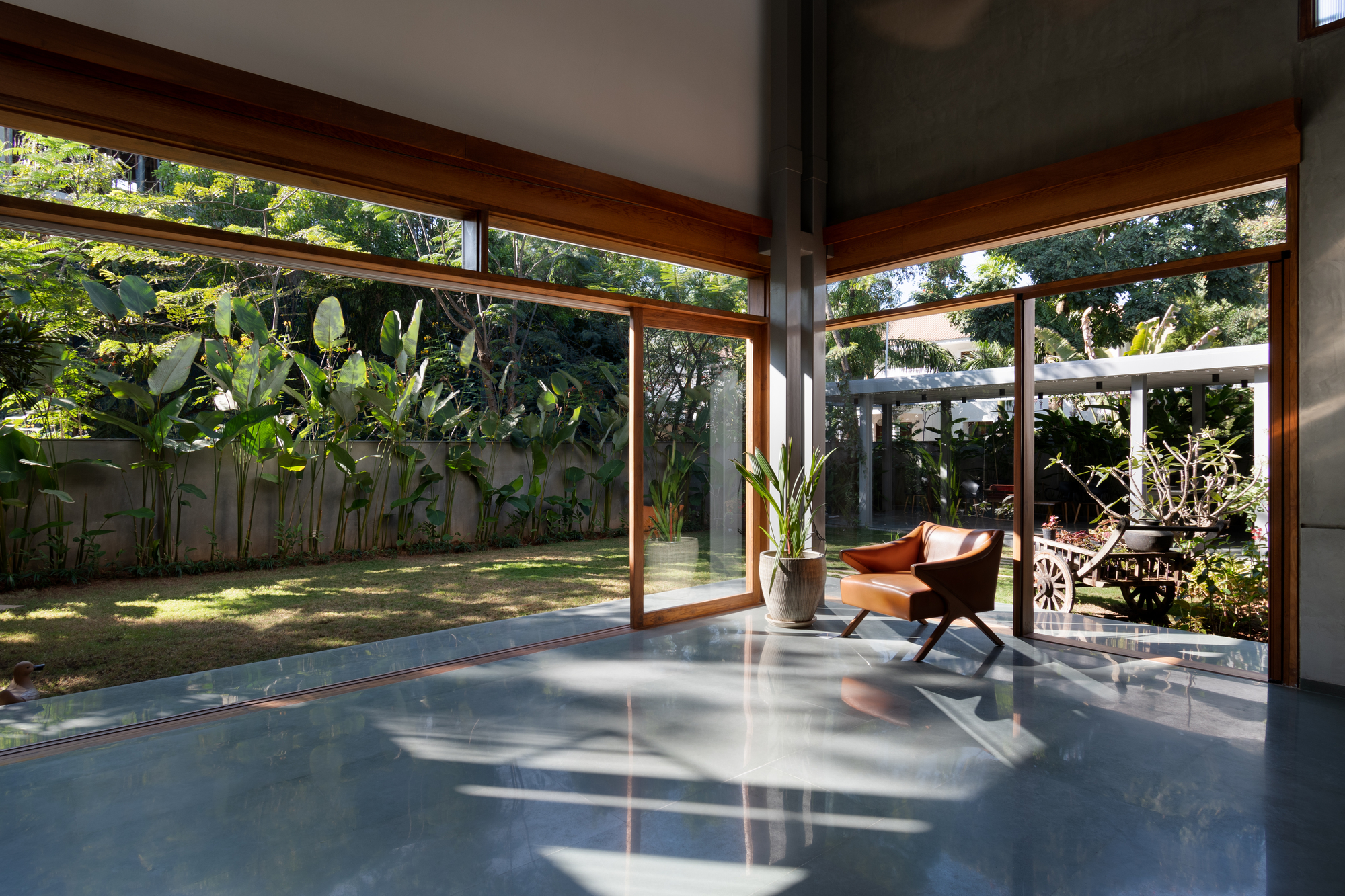 Glass-walled interior space with timber frames opening onto a grassed courtyard with banana plants