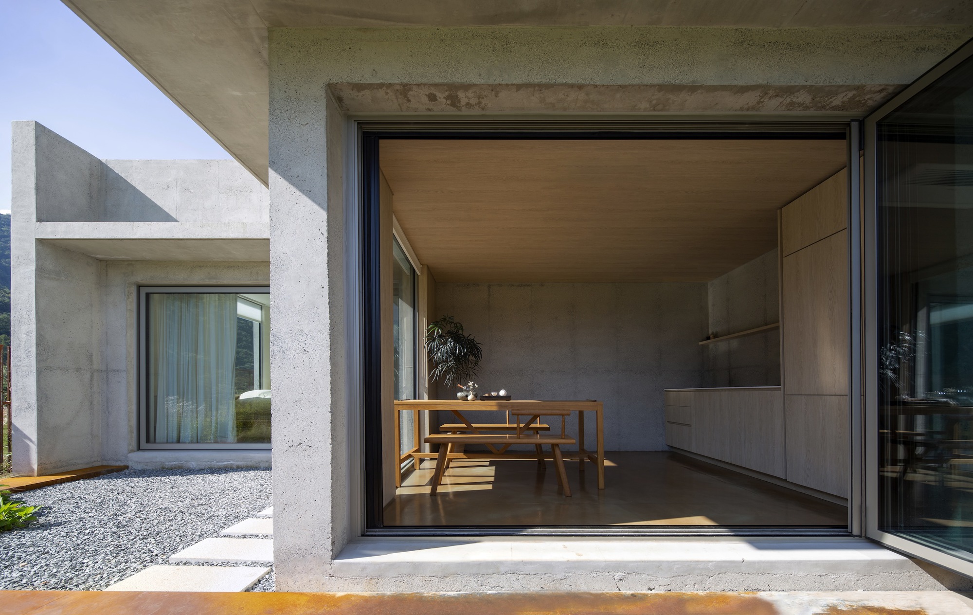 Glazed opening into interior dining area with wooden table and concrete-clad kitchen under afternoon sunlight