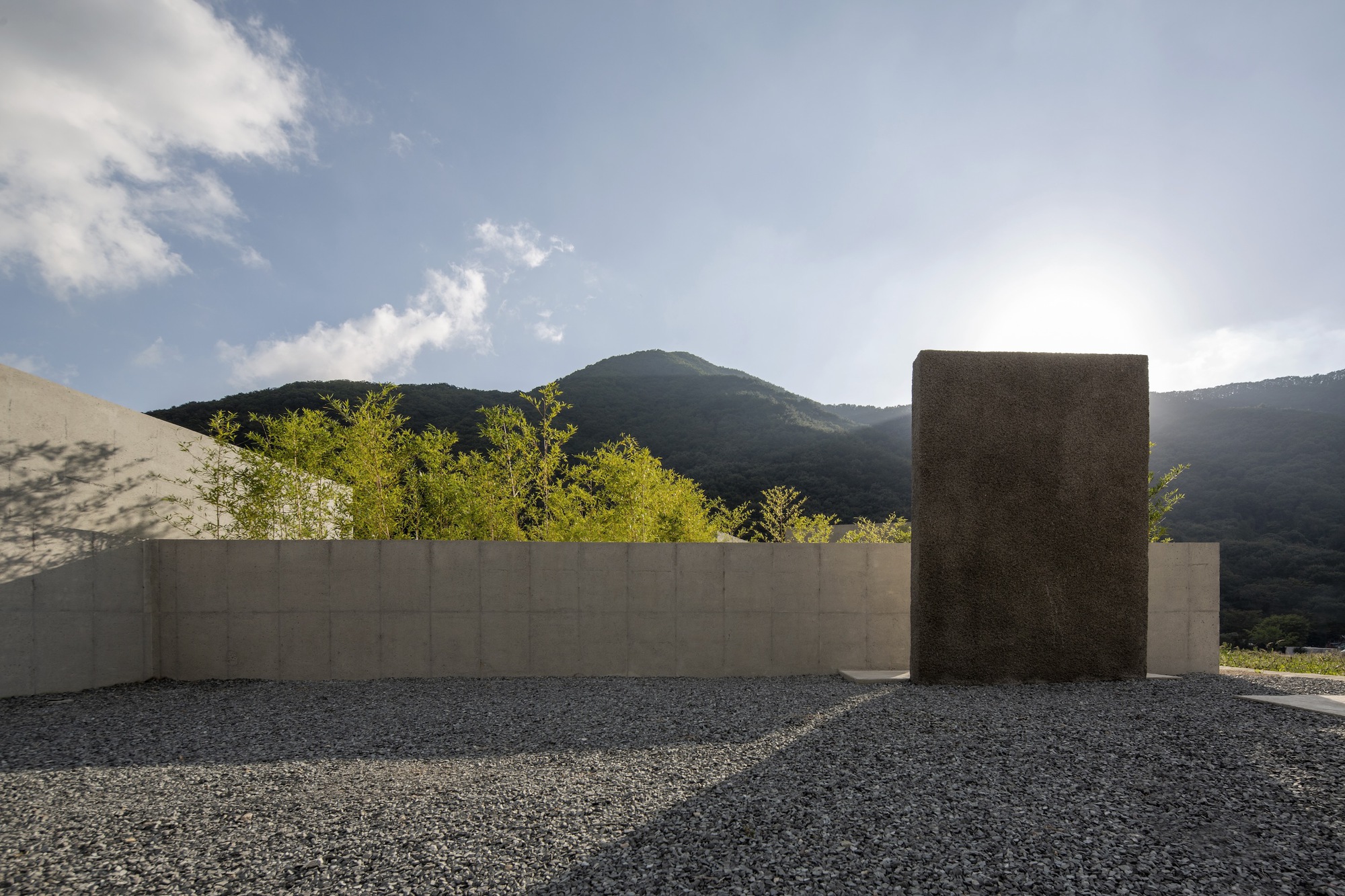Concrete perimeter wall with freestanding textured tower on gravel surface beneath hillside in late afternoon light