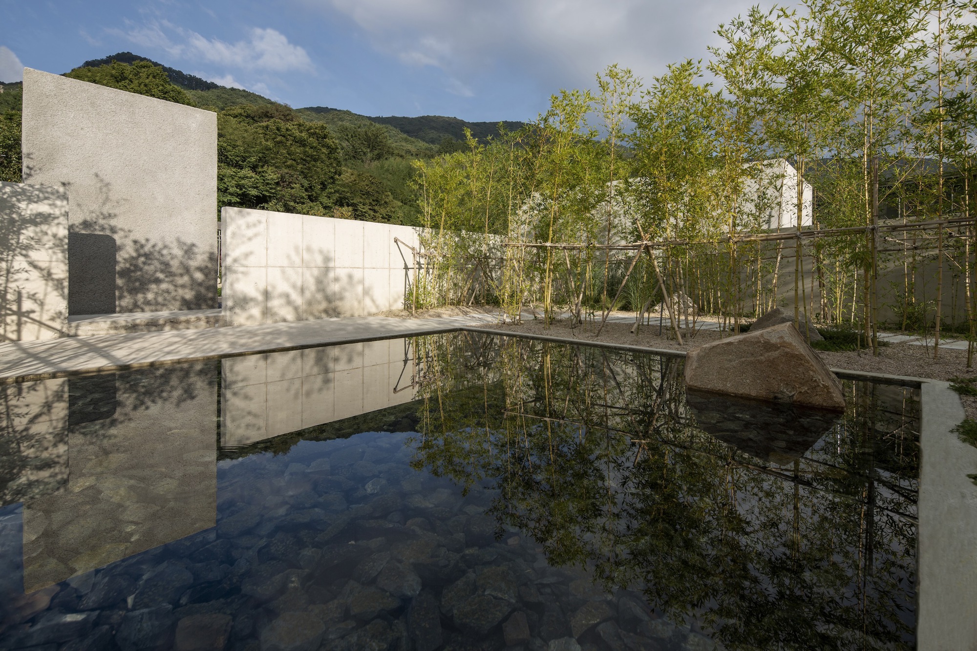 Reflecting pool mirroring white concrete walls and bamboo grove beneath clouded mountain slopes