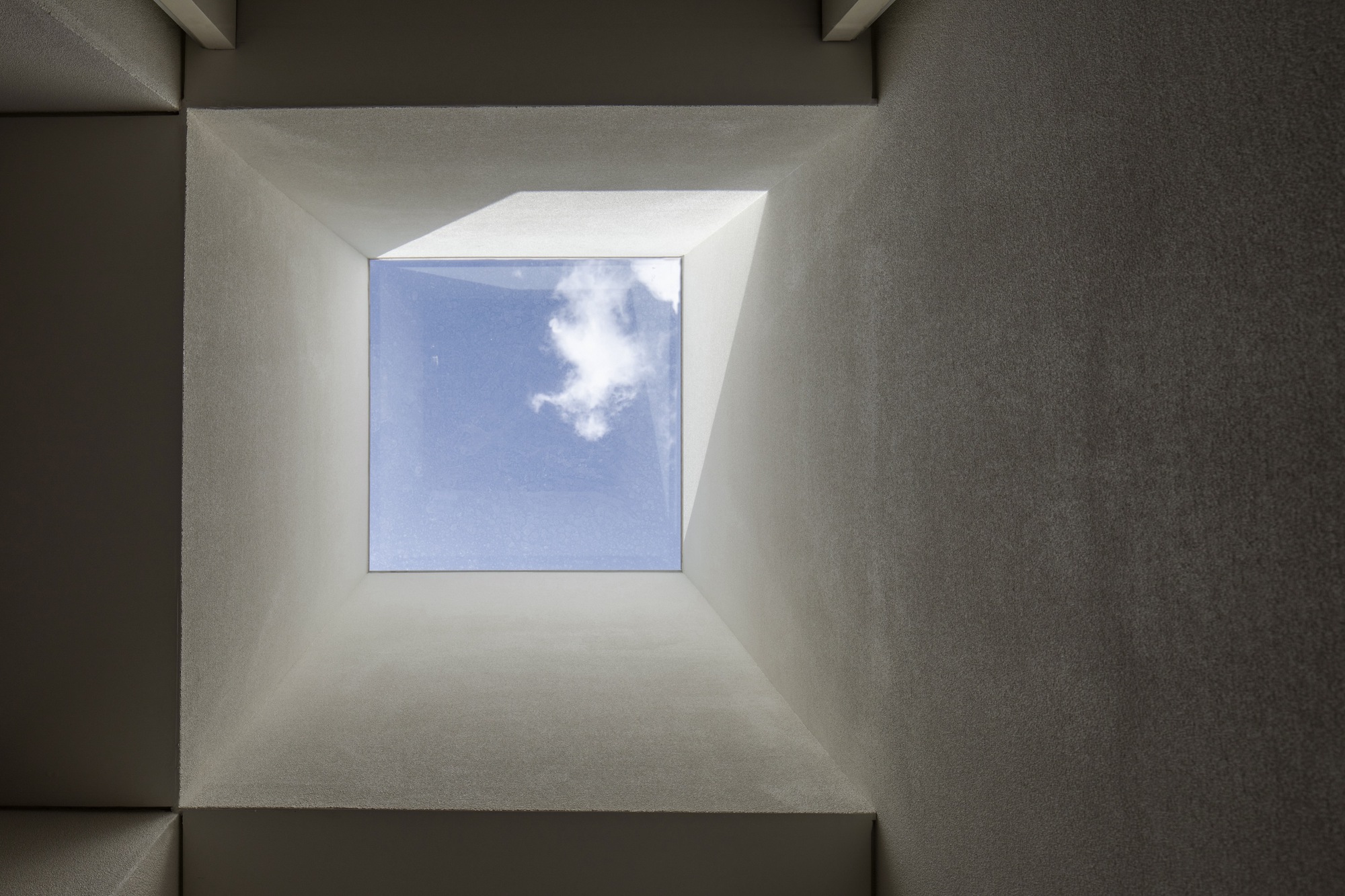 Upward view into a square skylight framed by sloped plaster walls with white clouds overhead