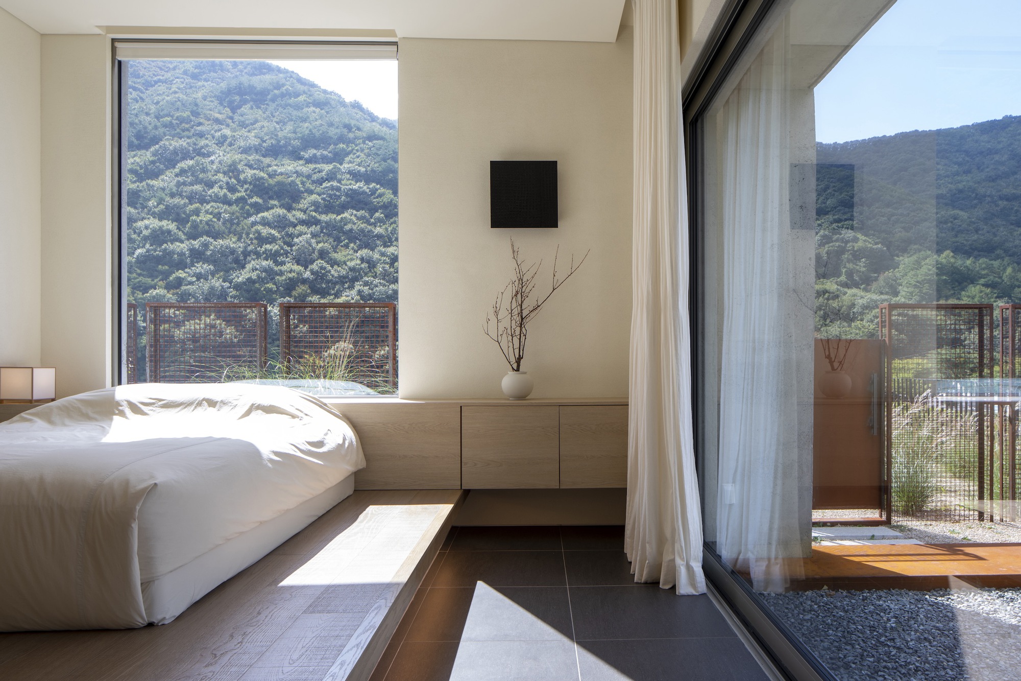 Bedroom corner with low platform bed and glass doors opening to a gravel courtyard in morning light