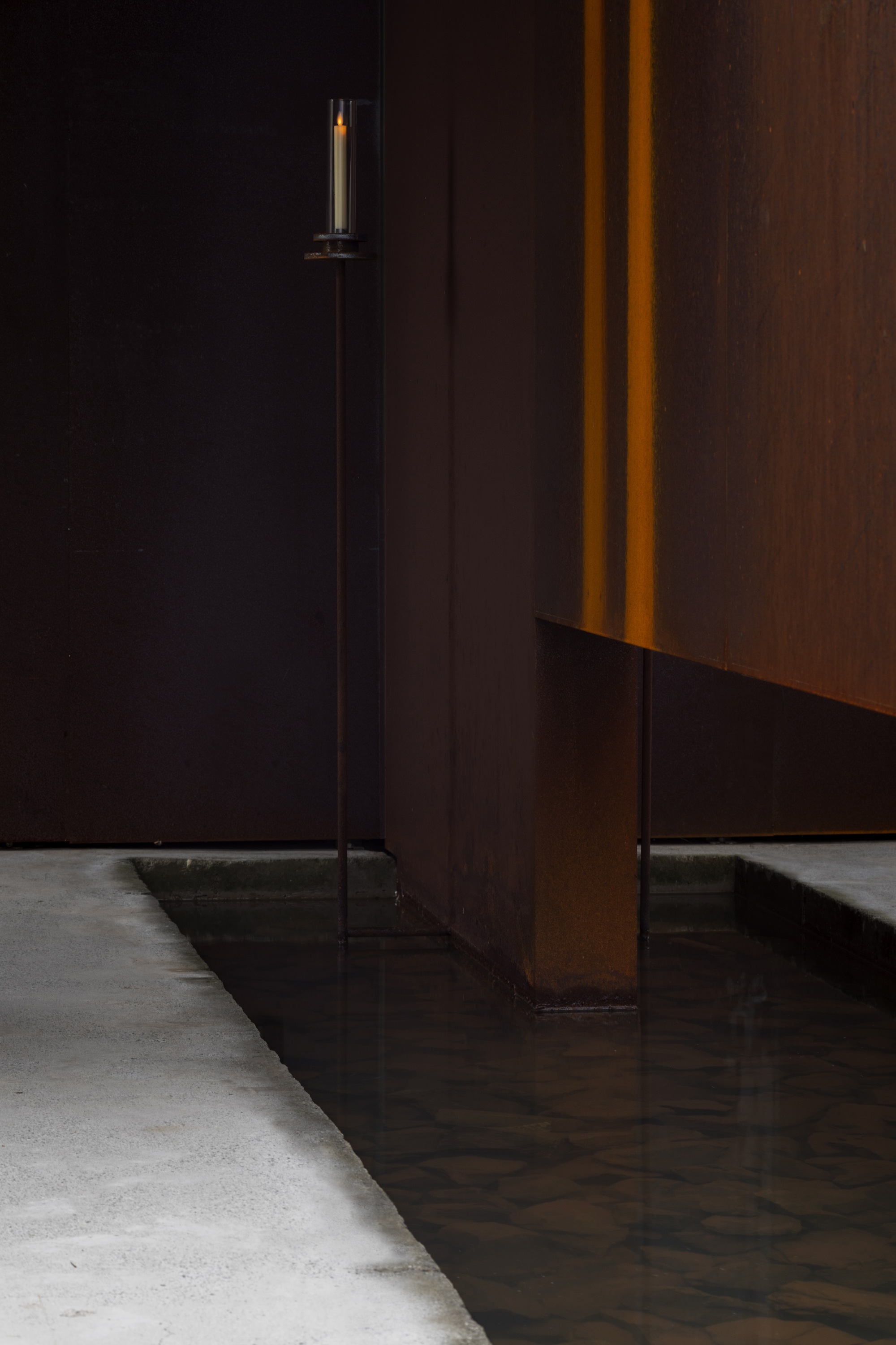 Detail of a weathered steel panel meeting a concrete floor with a tall candle in shadow