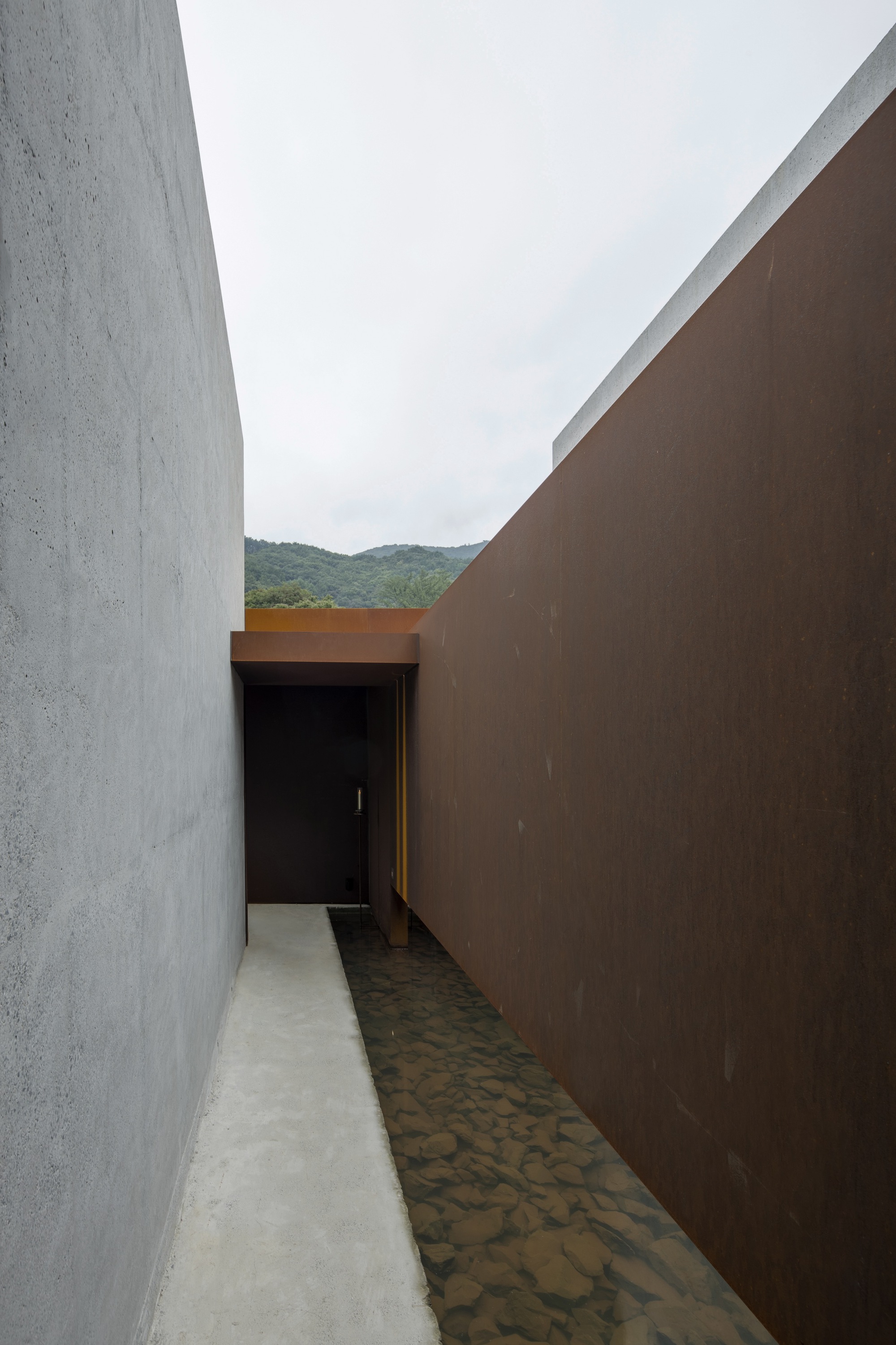 Narrow passage between concrete and weathered steel walls leading to a doorway under overcast sky