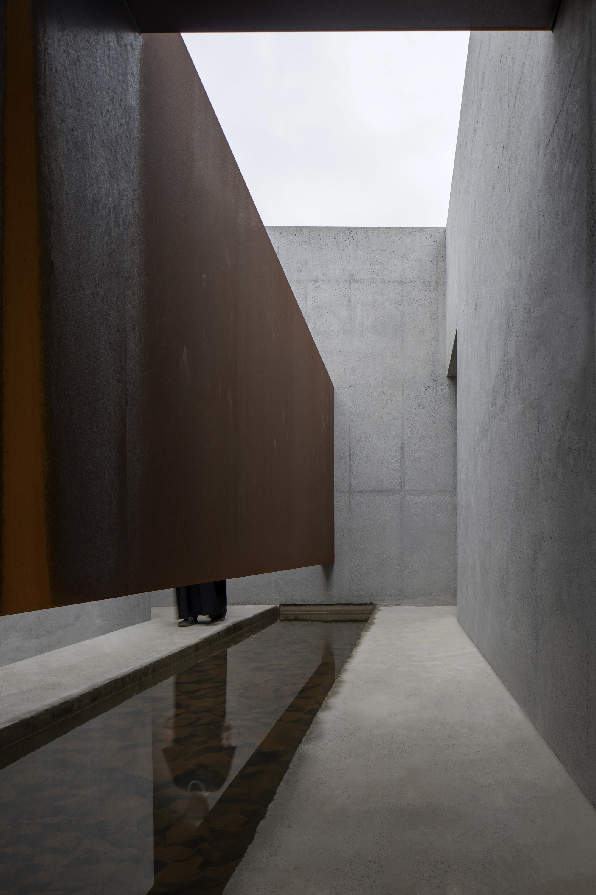Courtyard with shallow reflecting pool between concrete walls and angled weathered steel plane overhead