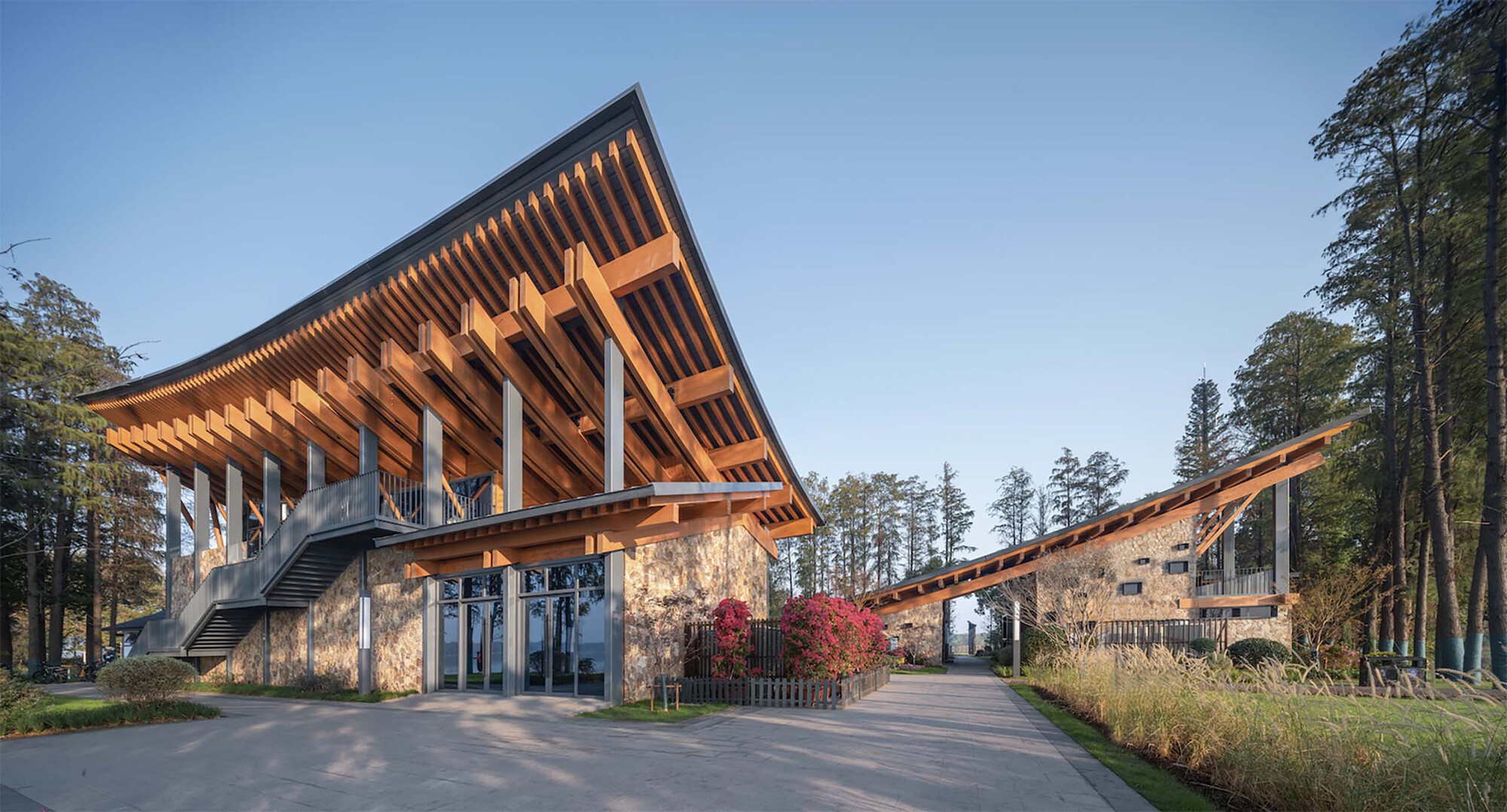 Entrance plaza with timber canopy sheltering stone-clad lower level and connecting walkway at dusk