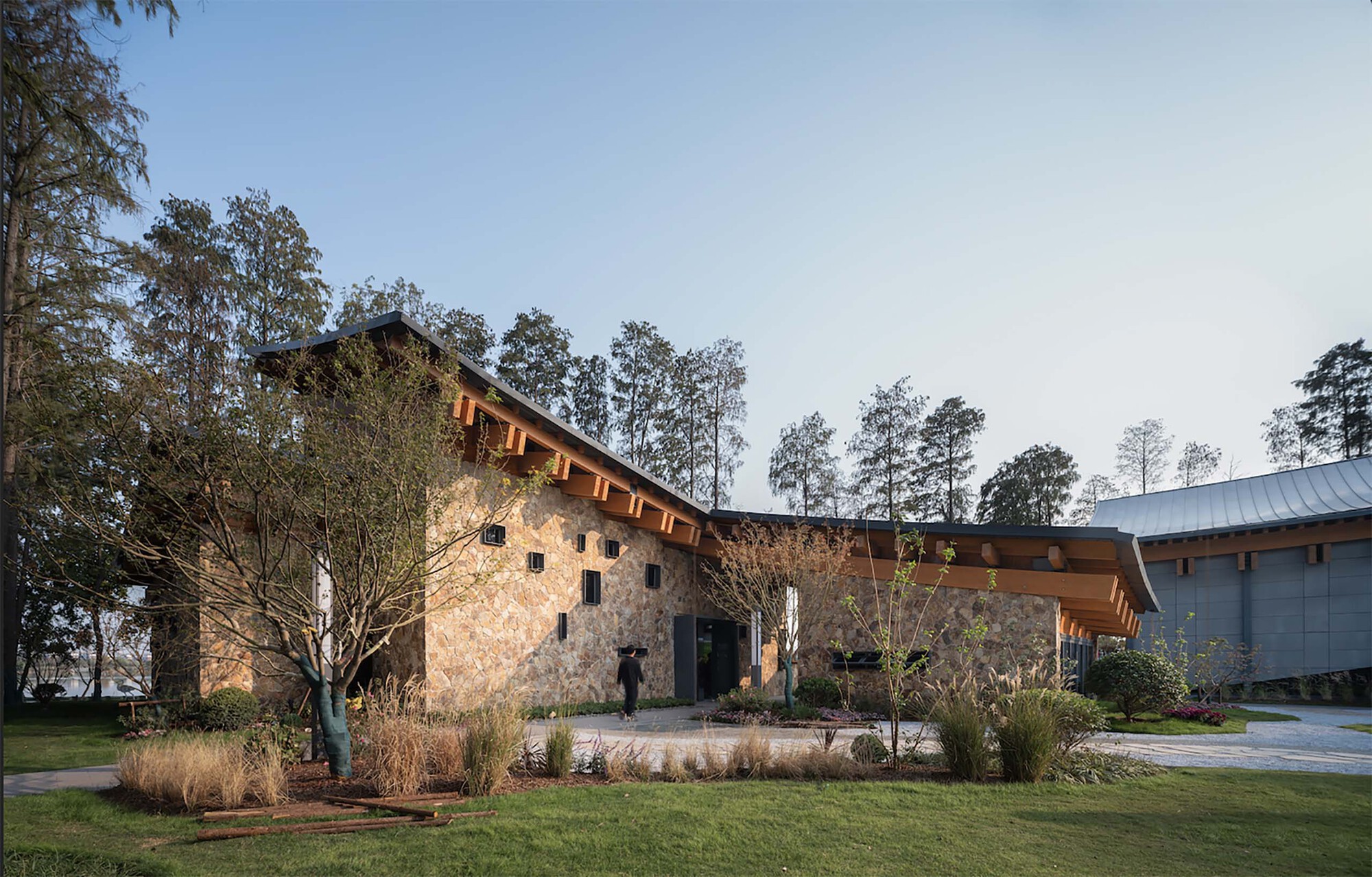 Rammed earth wall and planted beds with young trees set within mown lawn under pale sky
