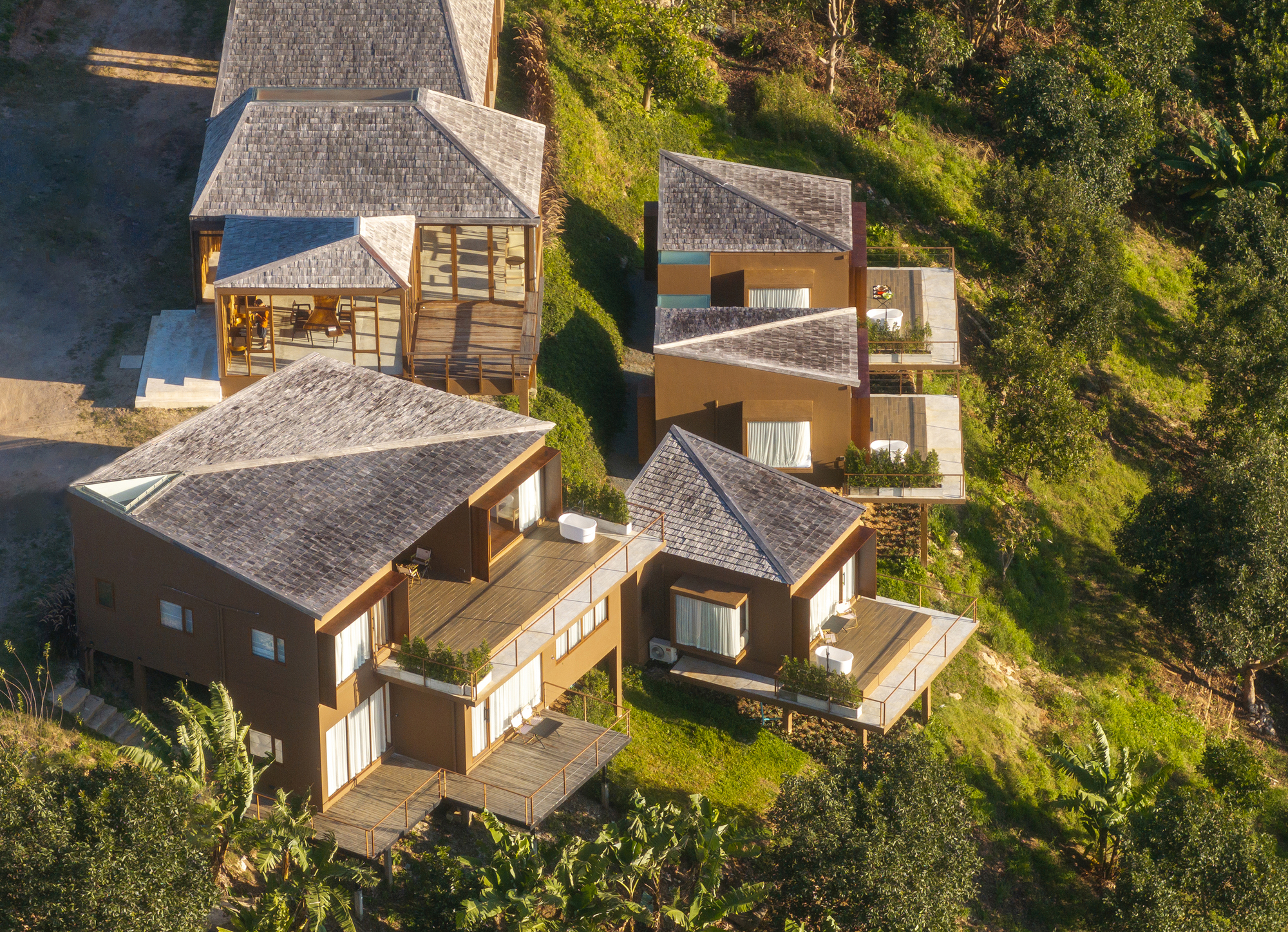 Aerial view of clustered gabled volumes with timber terraces embedded in green hillside vegetation