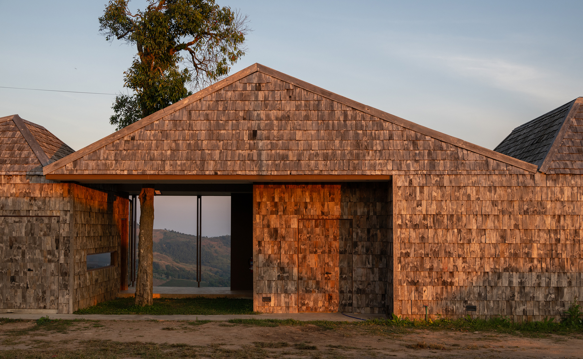 Gable-roofed volume clad in timber shingles with a central passageway framing distant hills at dusk